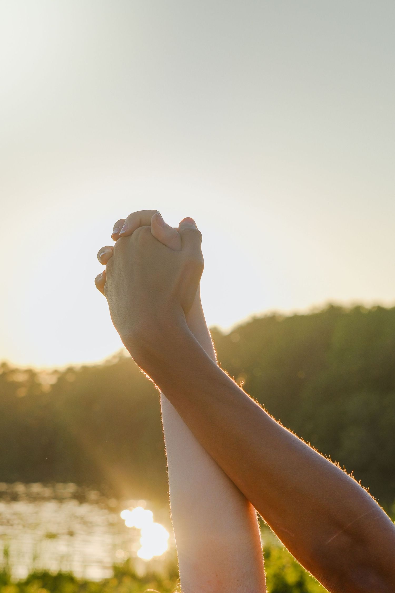 Two hands clasped together raised against a golden sunset sky with trees and water in the background.
