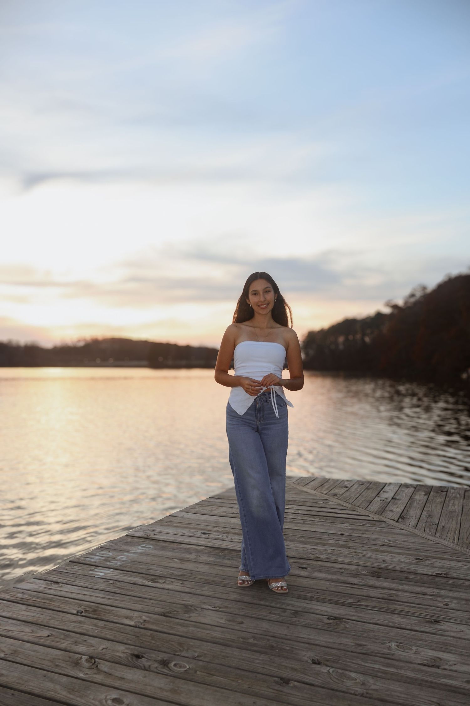 Young woman in white strapless top and wide-leg jeans standing on wooden dock at golden hour sunset.