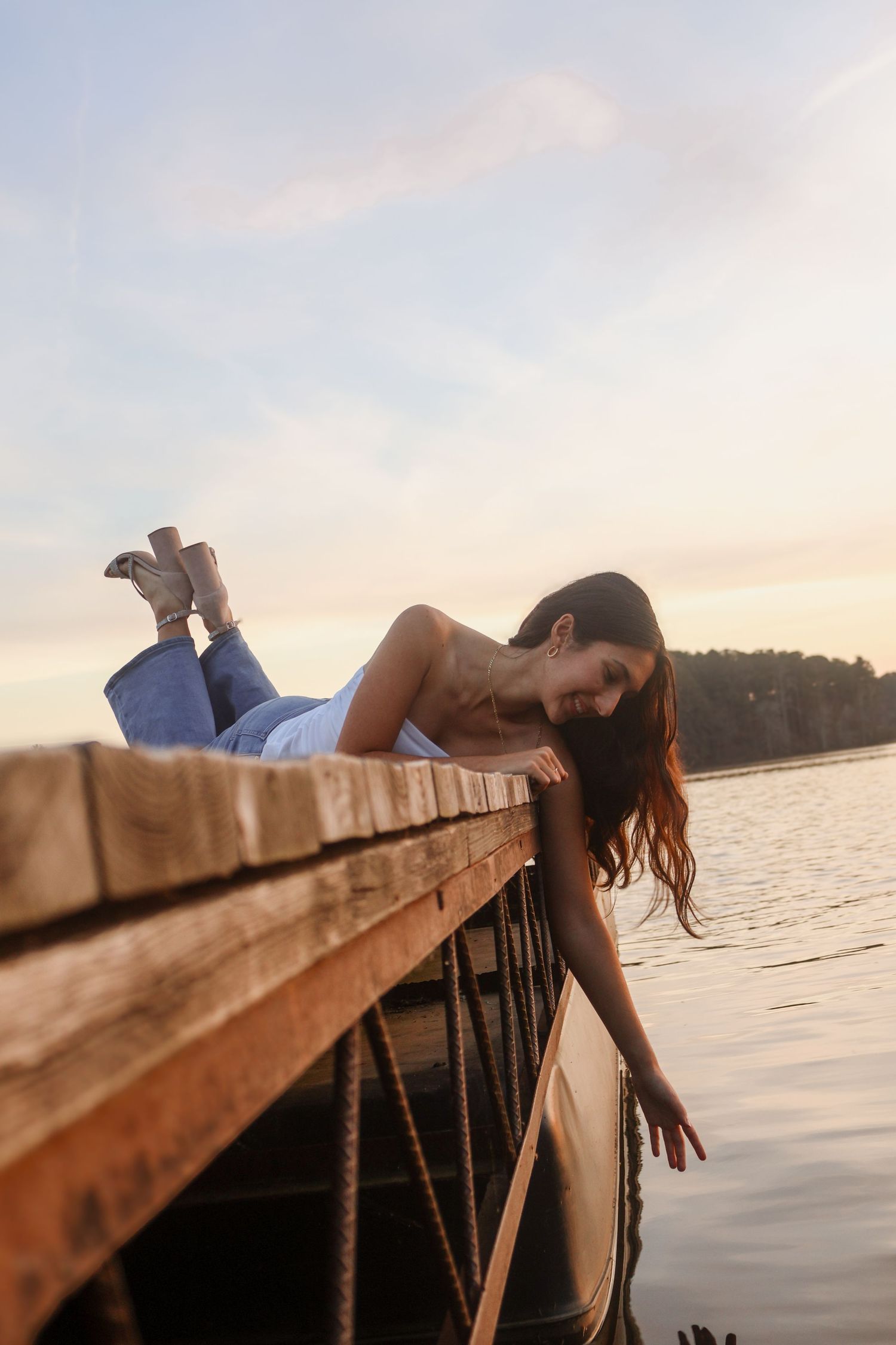 Young woman in jeans and boots lying on a wooden dock overlooking a calm lake at sunset.