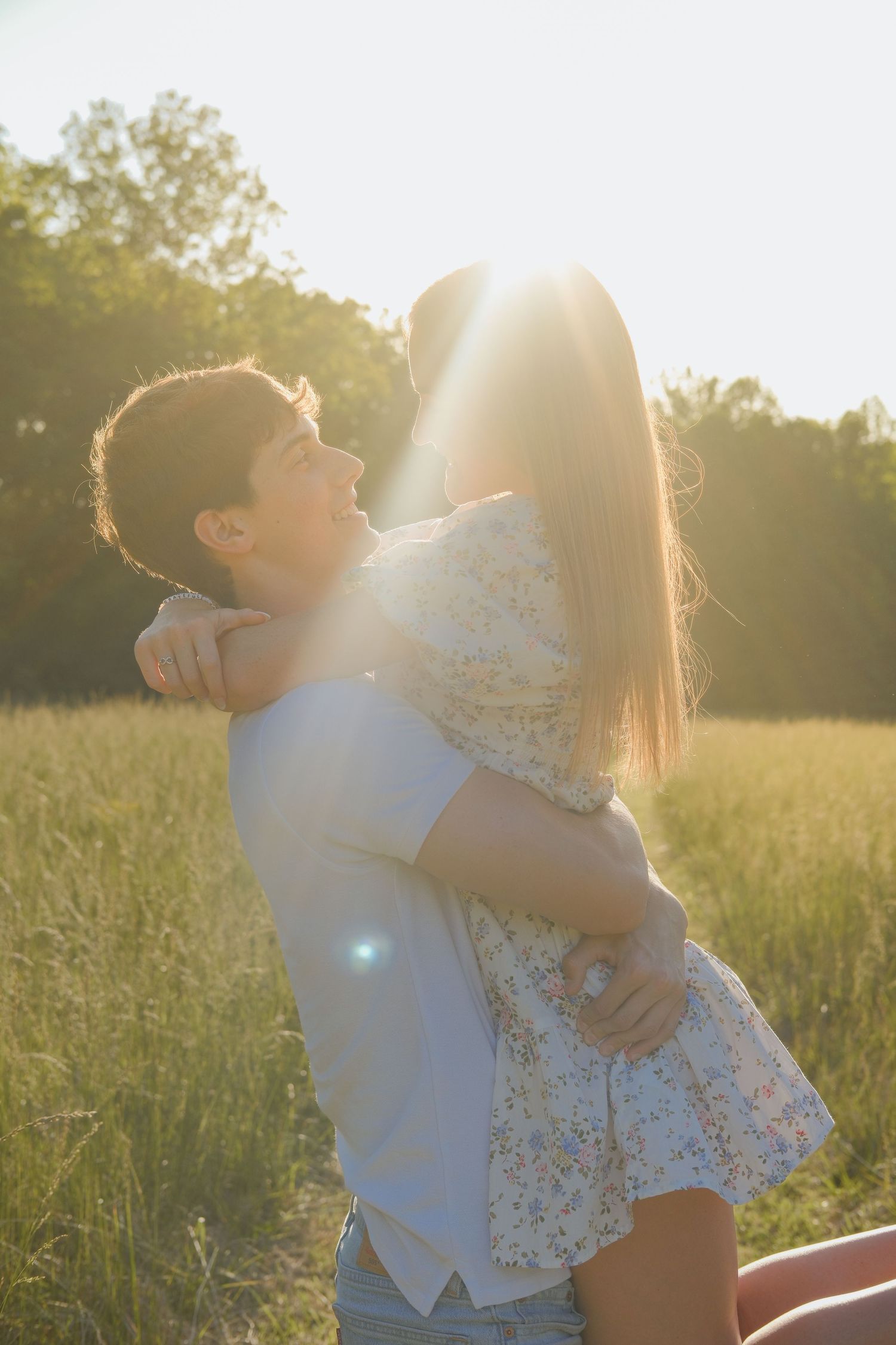 Couple embracing in a sunlit field, golden backlight glowing behind them, surrounded by green grass and trees.
