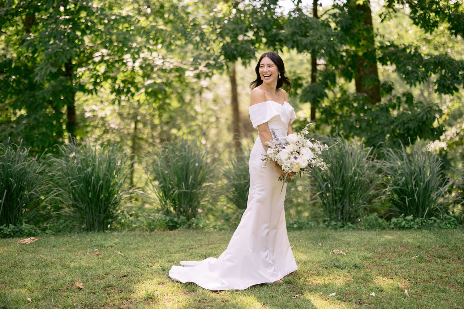 Bride in flowing white dress walks through sunlit garden setting with natural greenery.