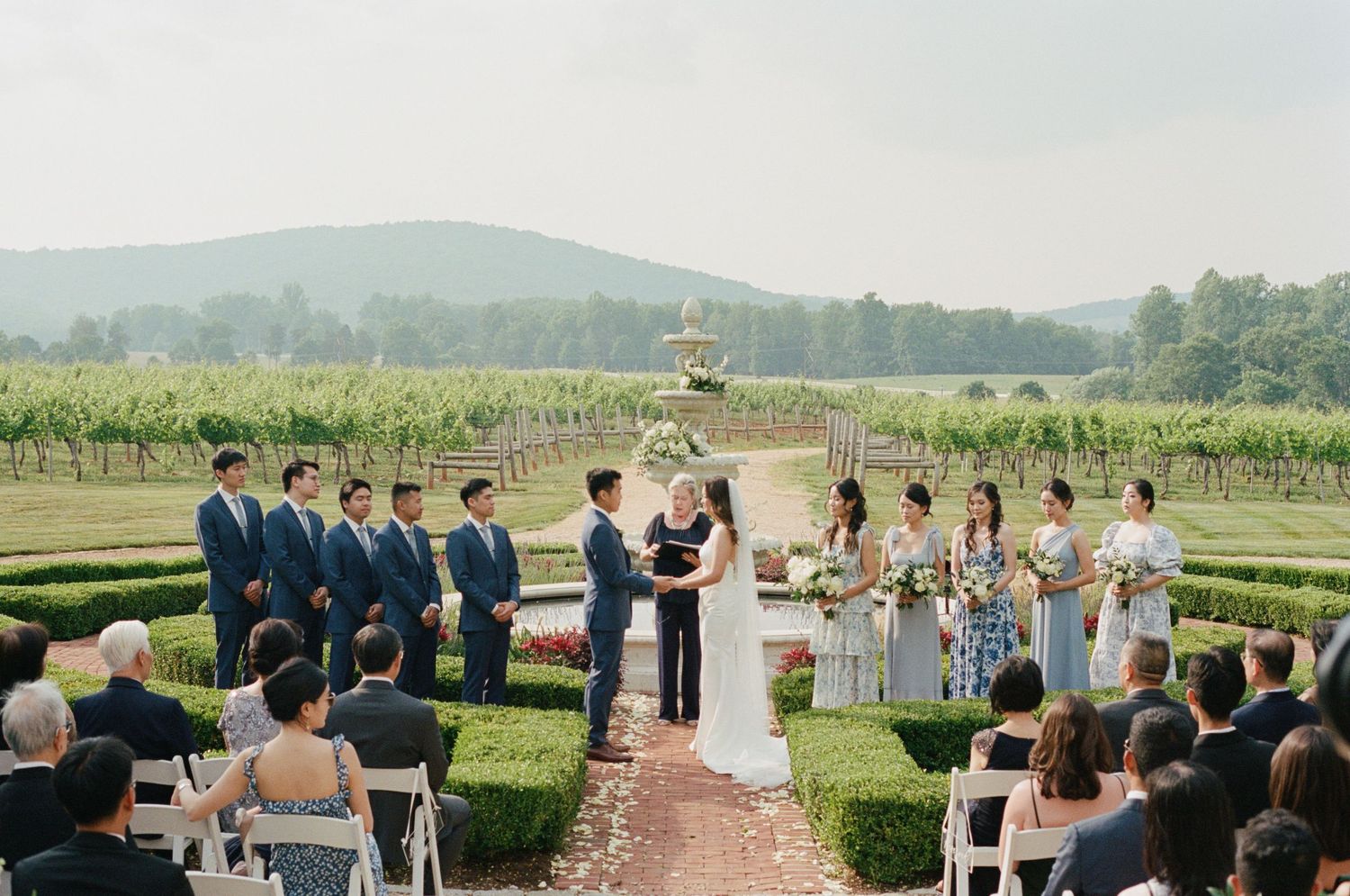 Outdoor vineyard wedding ceremony with guests seated in rows facing scenic landscape.