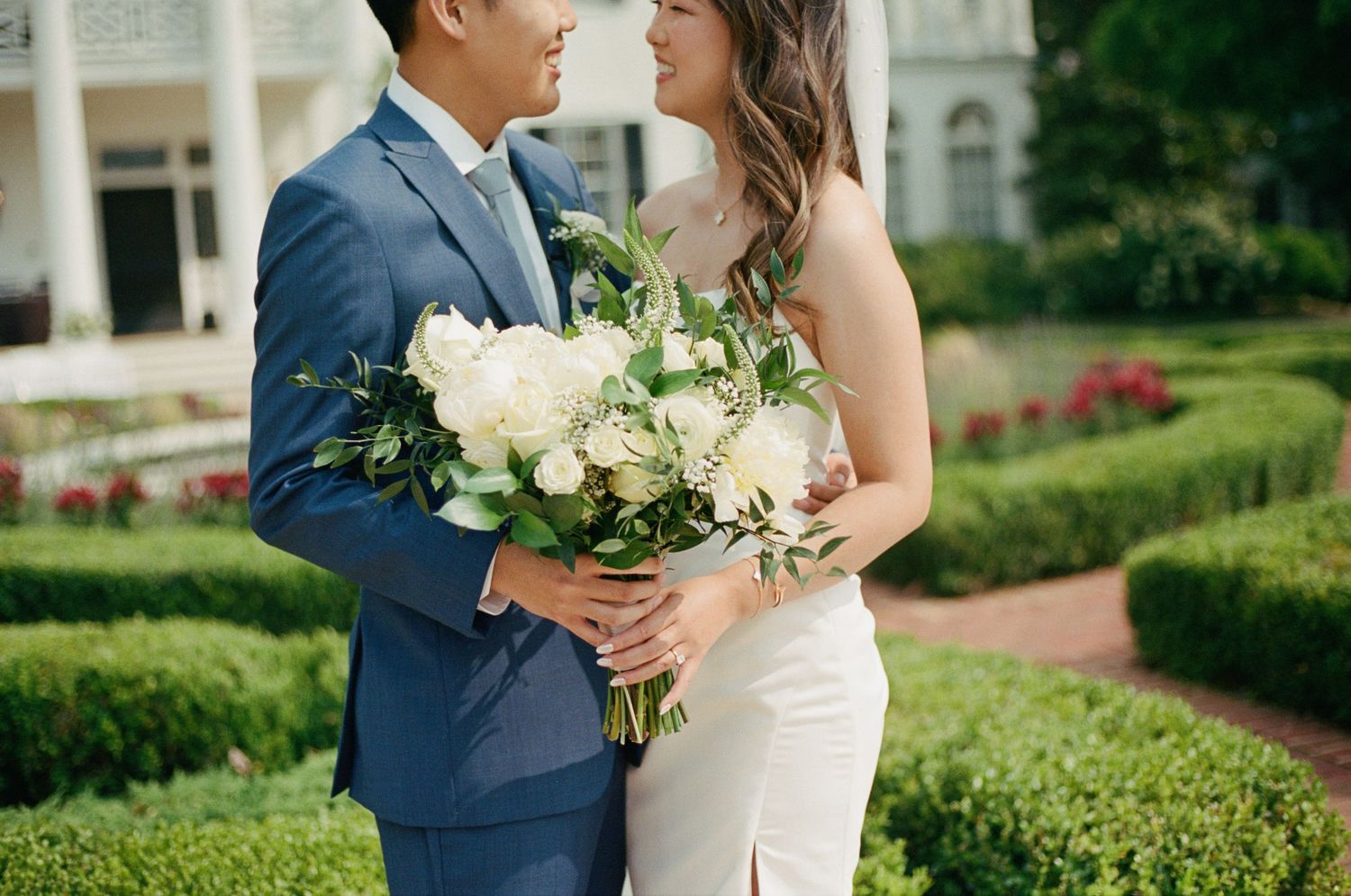 Elegant wedding couple posing in a manicured garden with white floral bouquet.