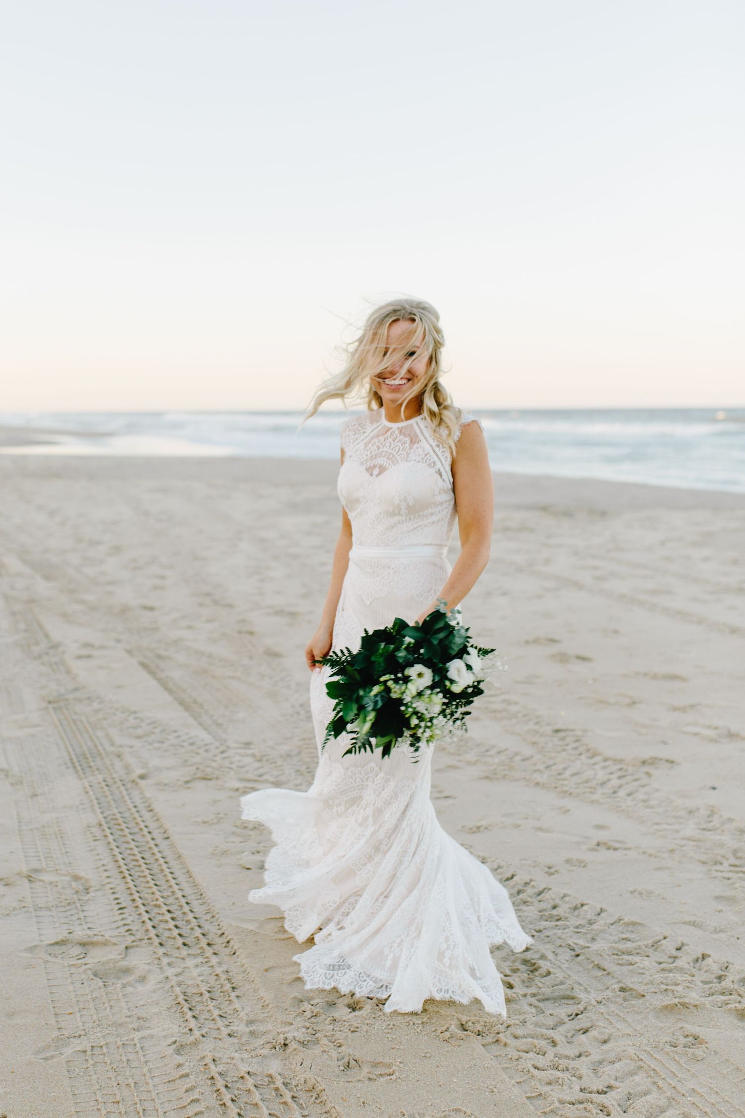 Bride in lace dress poses on sandy beach with flowing bouquet.