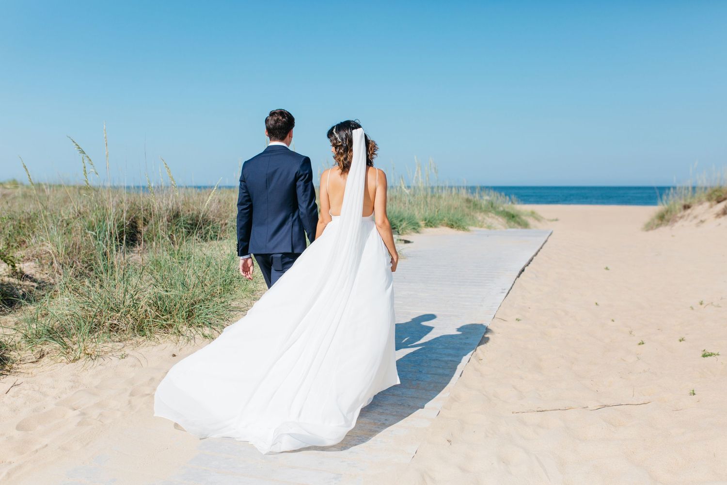 A couple walks along a sandy beach path with ocean views on their wedding day.