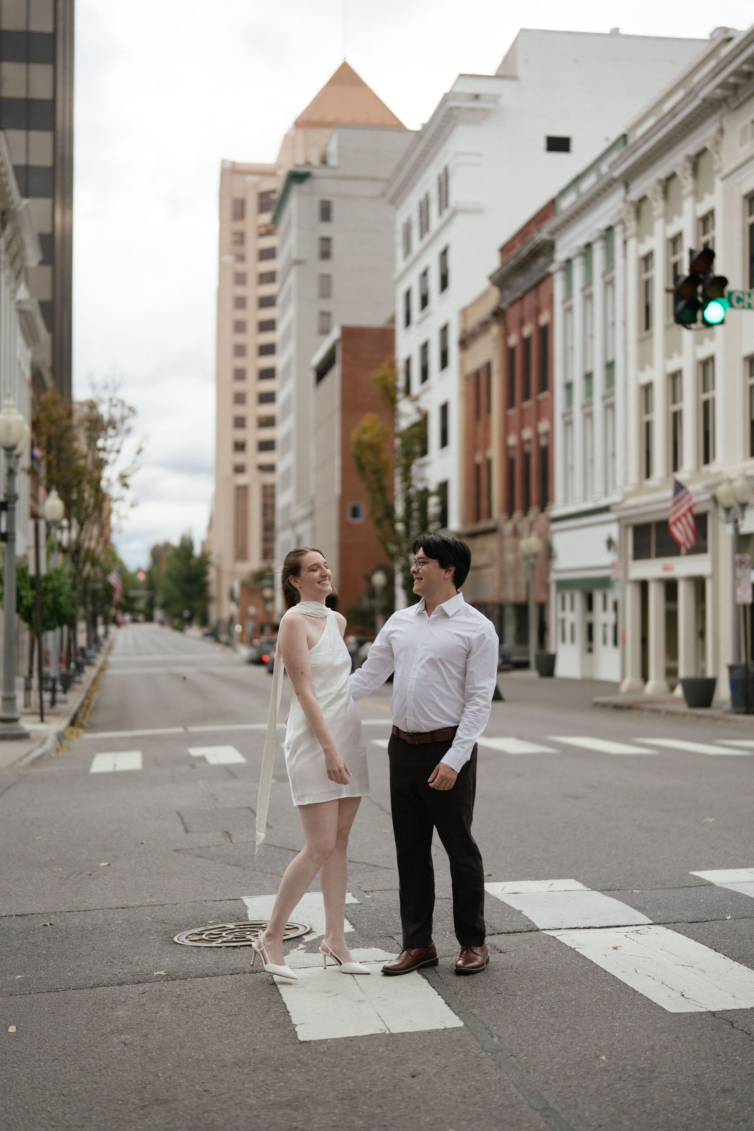 A couple stands together on a crosswalk in an urban setting with tall buildings and historic architecture in the background.