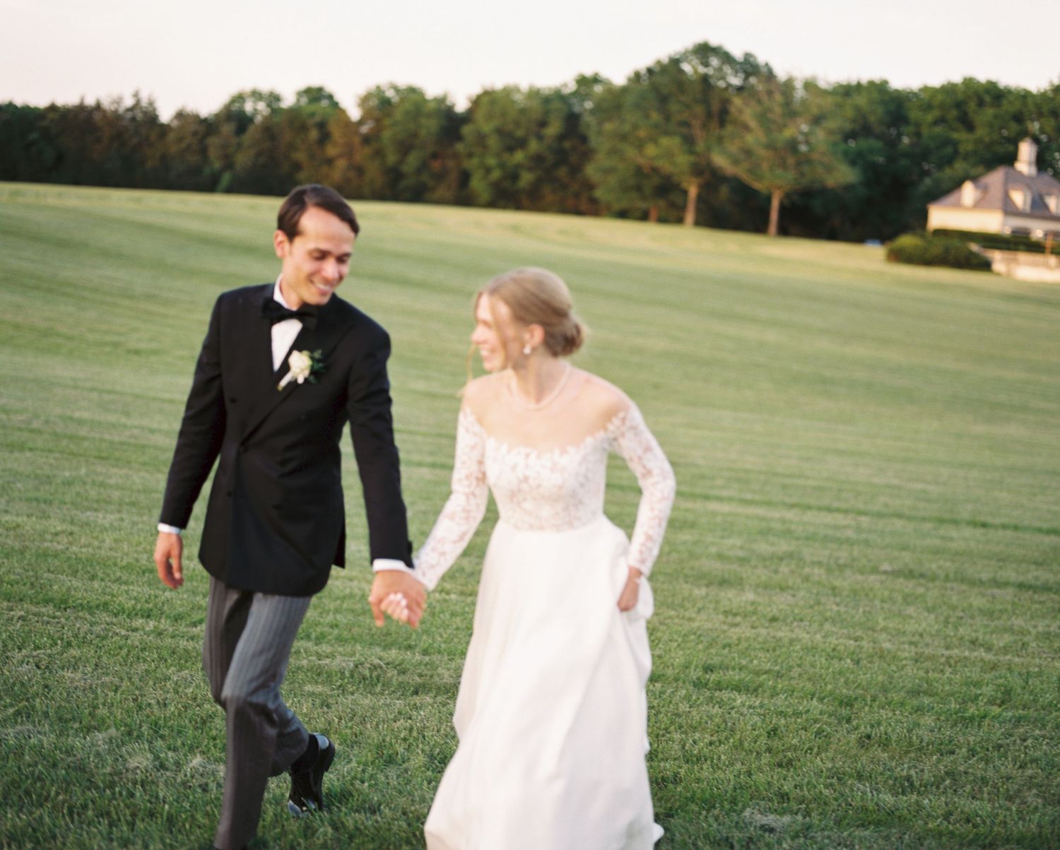 Couple running playfully across a green field in wedding attire at sunset.