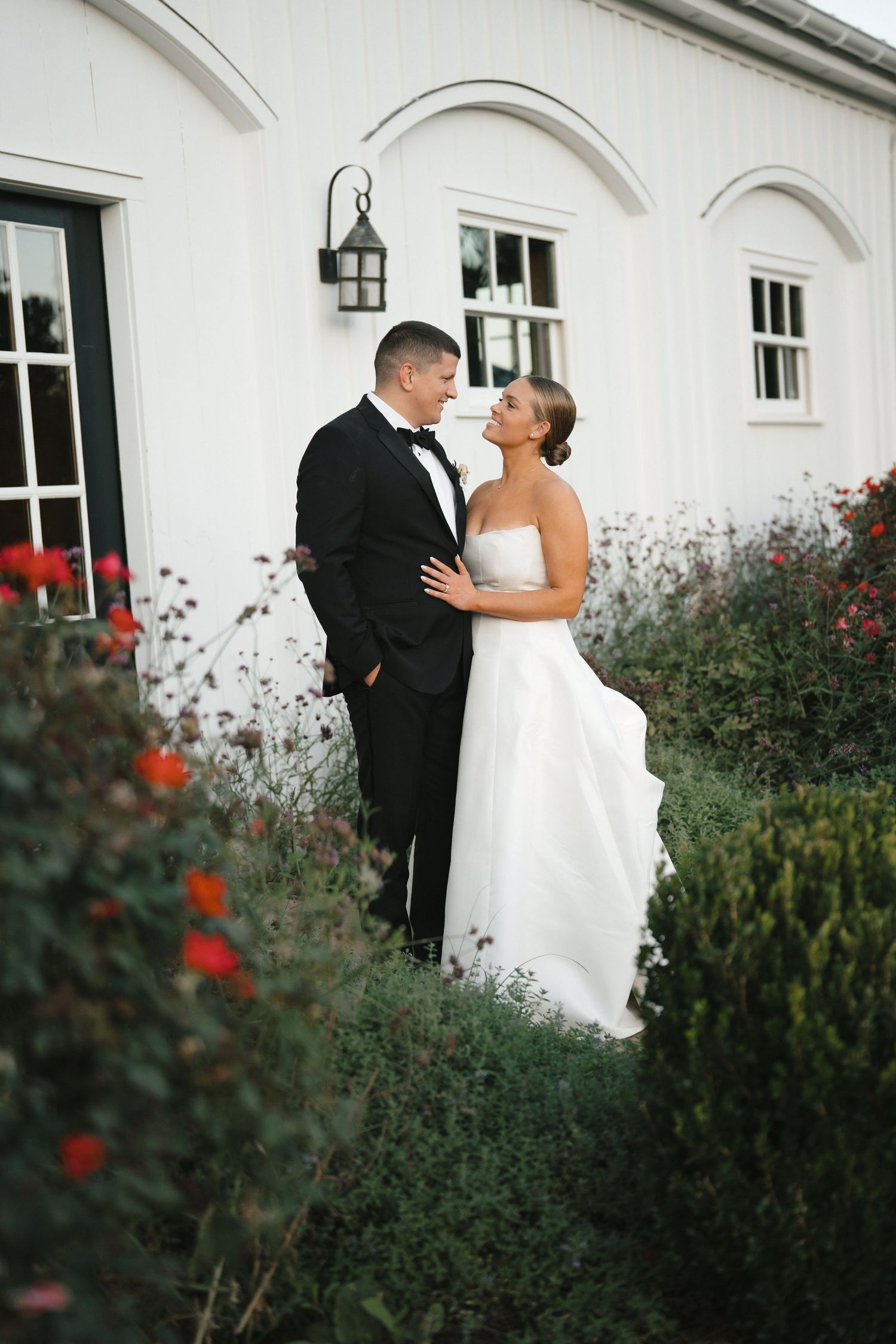 A couple in wedding attire embrace in a garden with red flowers and a white building facade in the background.