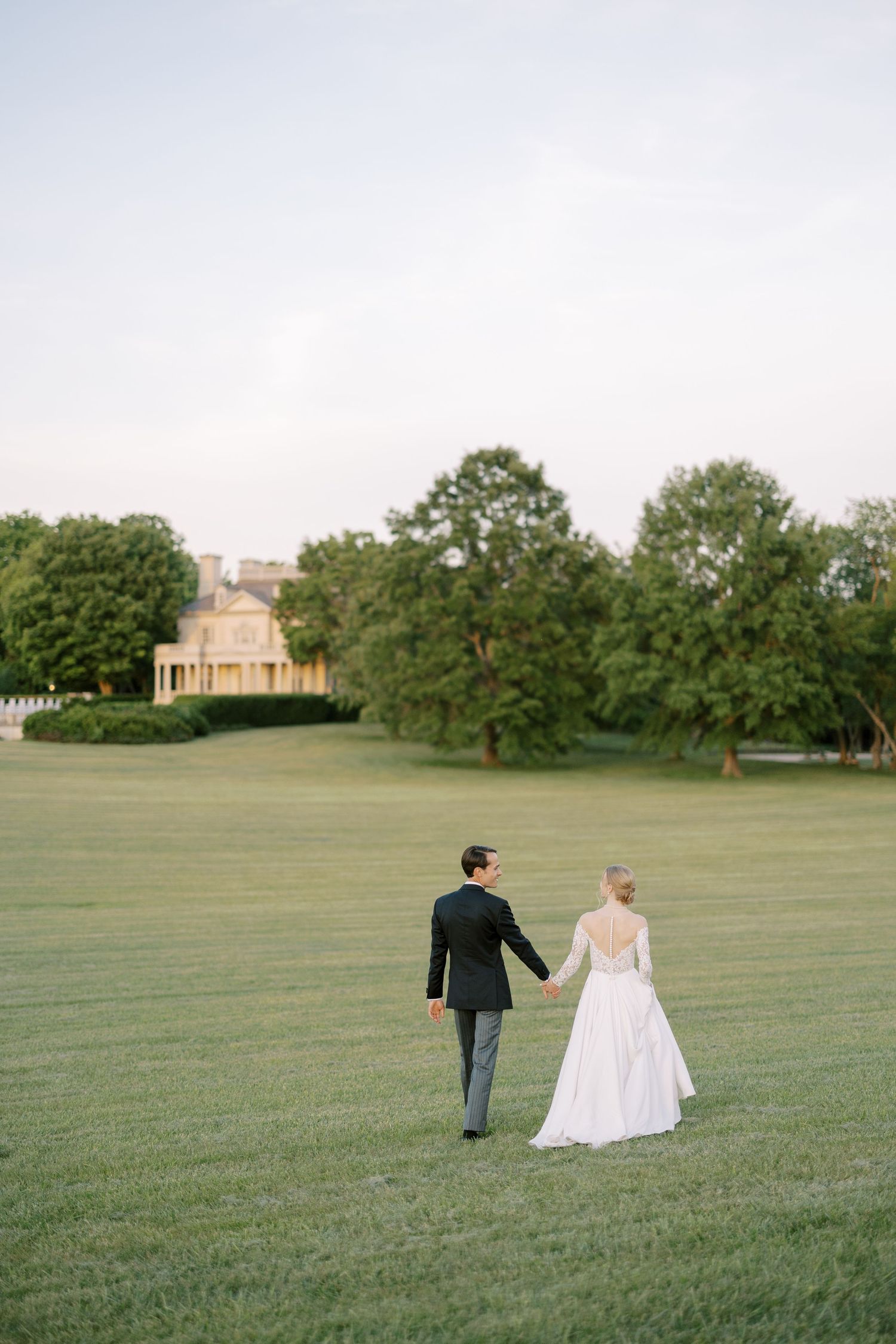 A couple walks hand in hand across a vast green lawn on their wedding day.