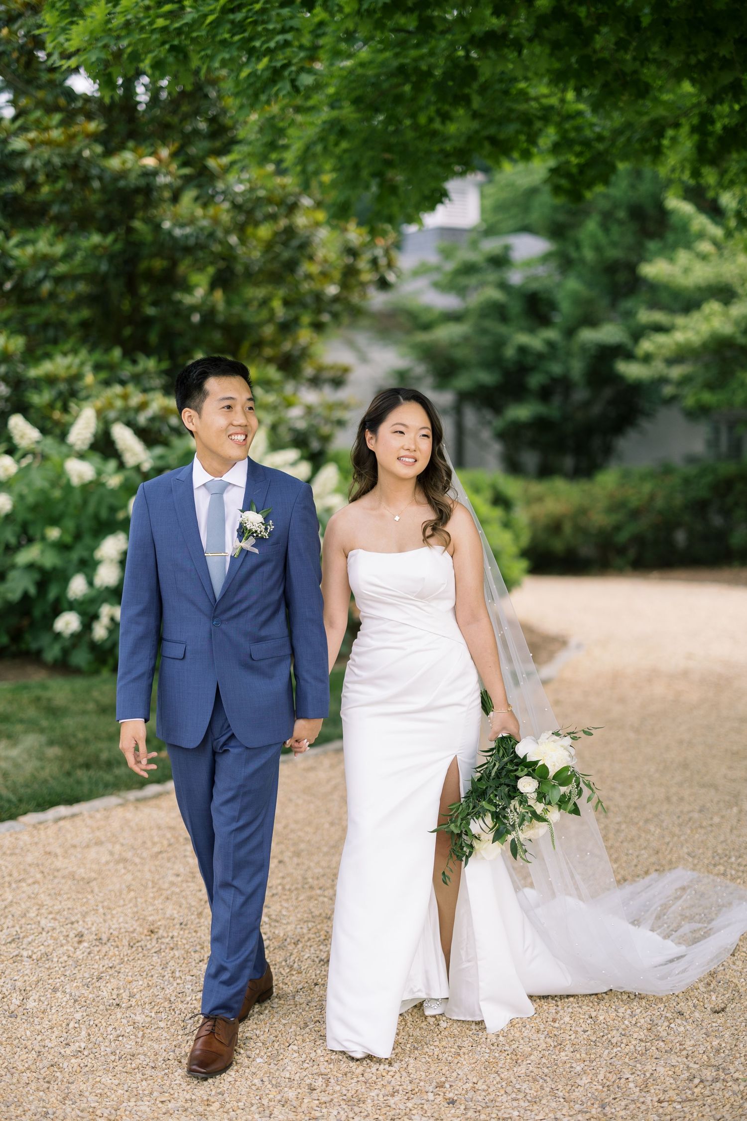 A couple walks along a gravel path surrounded by lush greenery on their wedding day.