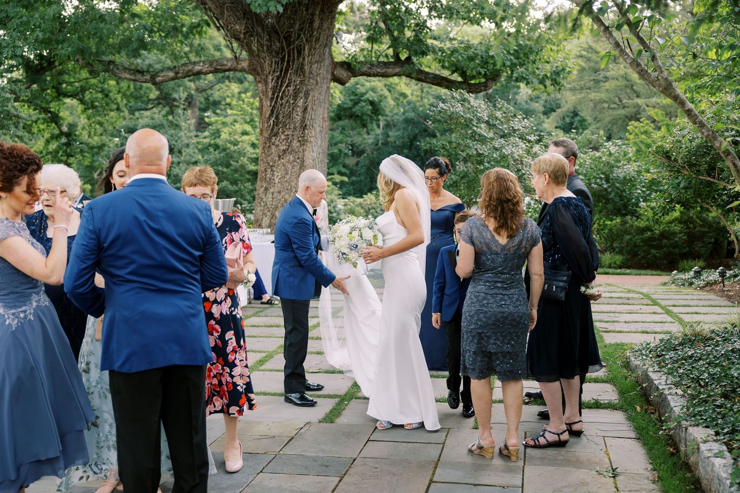 Wedding guests gather outdoors under trees for a celebration.