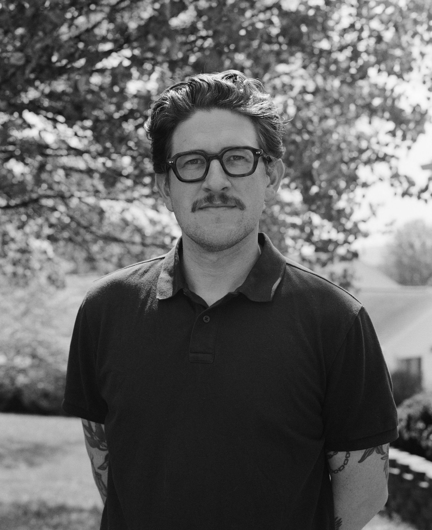 Black and white portrait of a man with glasses and mustache, tattooed arms, standing outdoors under trees.