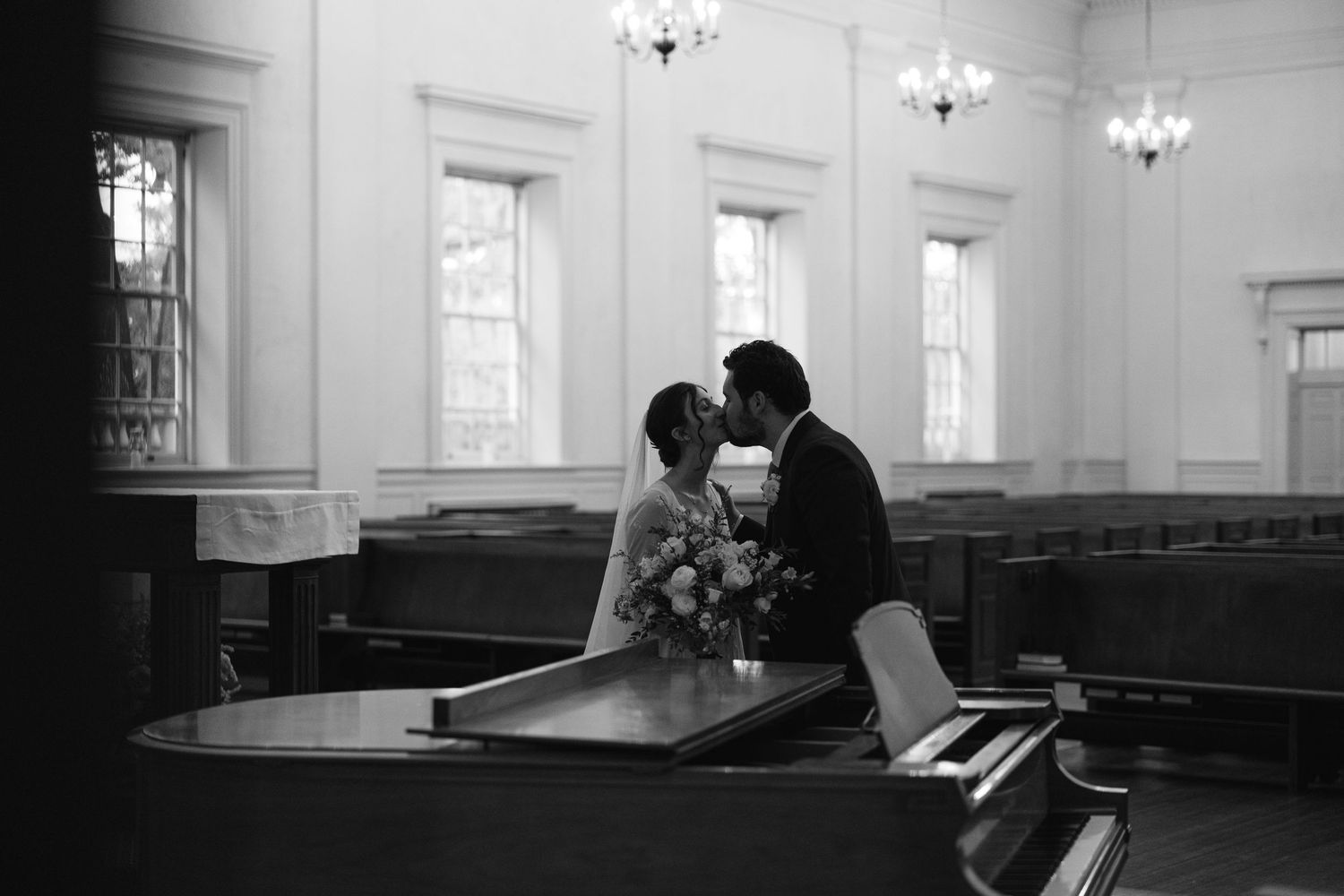 A romantic couple shares an intimate moment while dancing in an elegant ballroom with chandeliers and windows.