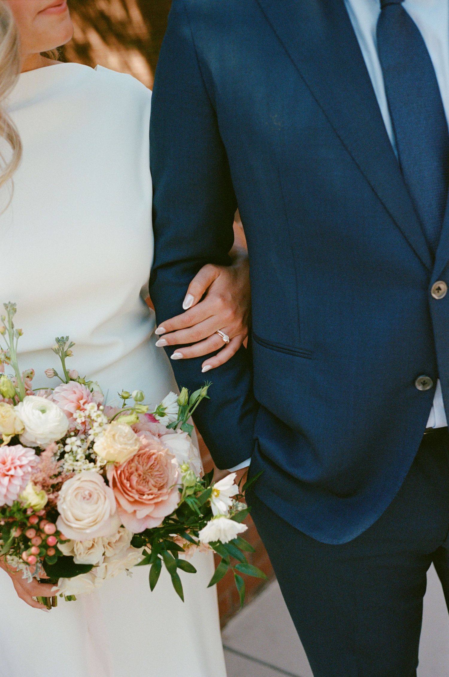 Close-up of couple holding hands at wedding with pink and white rose bouquet and navy suit.