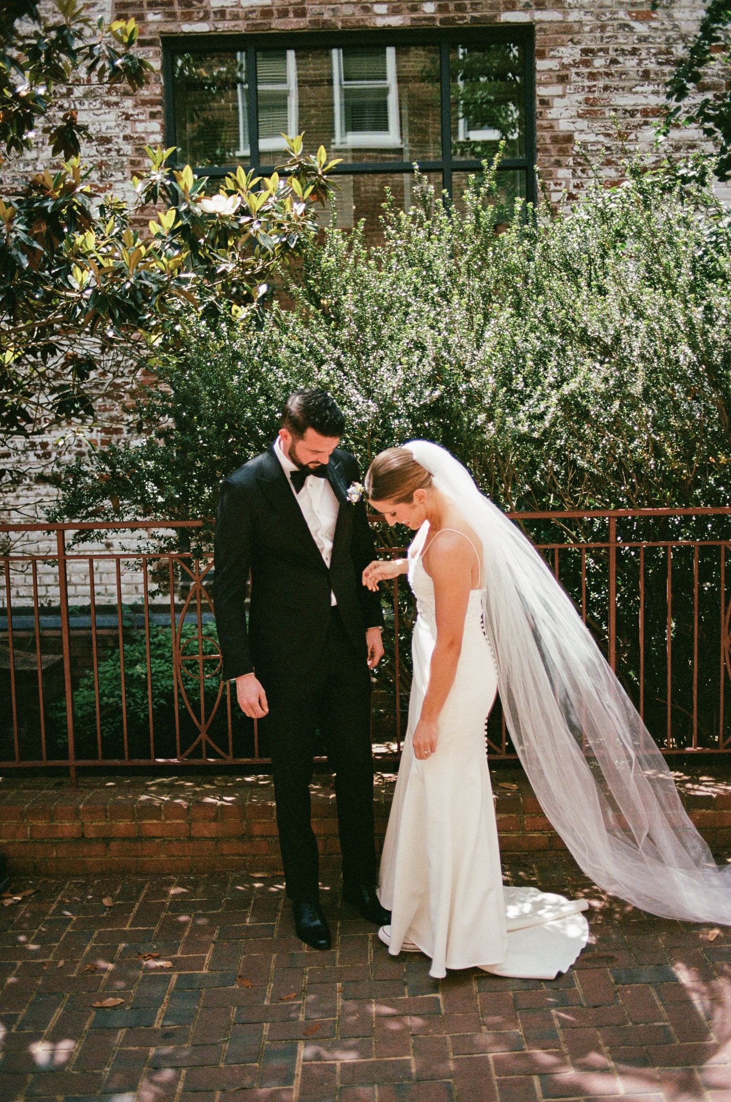Bride in flowing dress and veil poses against rustic brick wall with garden flowers.