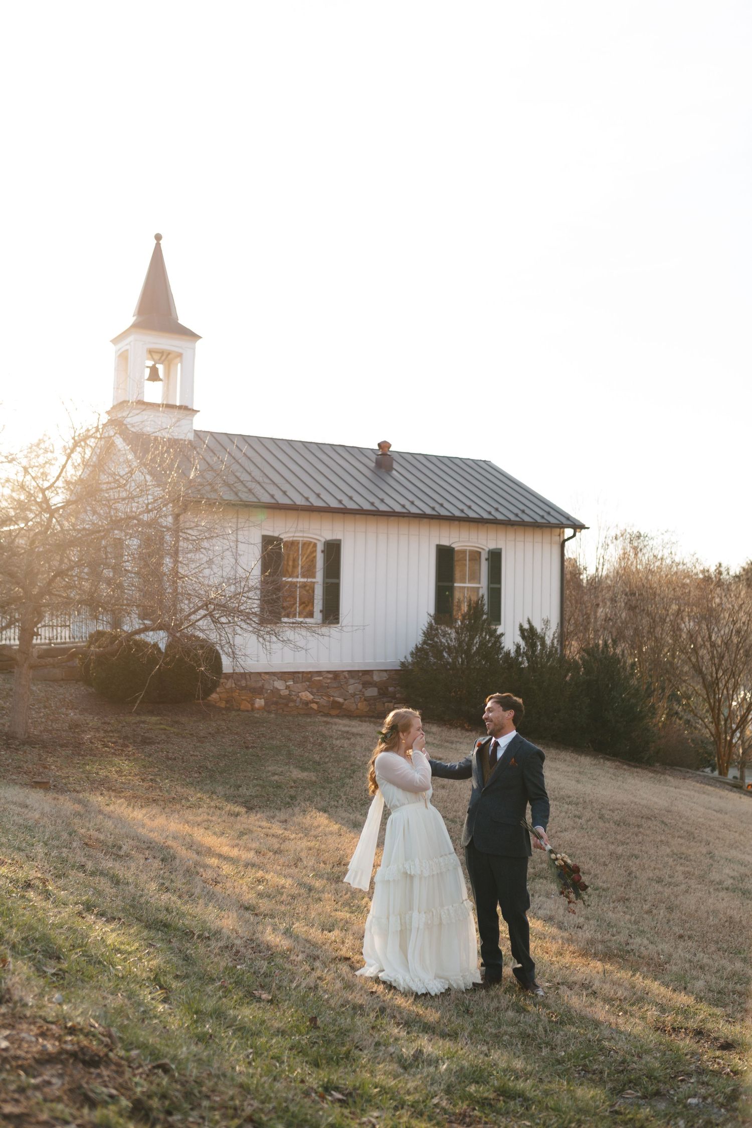 Wedding couple walks near white chapel during golden hour.