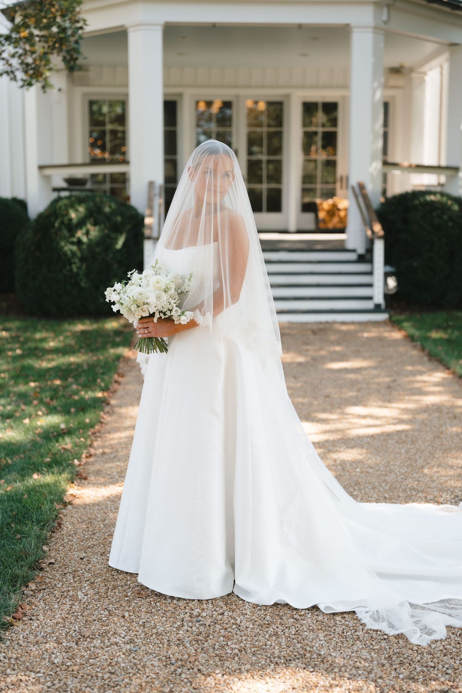 A bride in a flowing white wedding dress and veil stands in front of a classic white columned mansion.