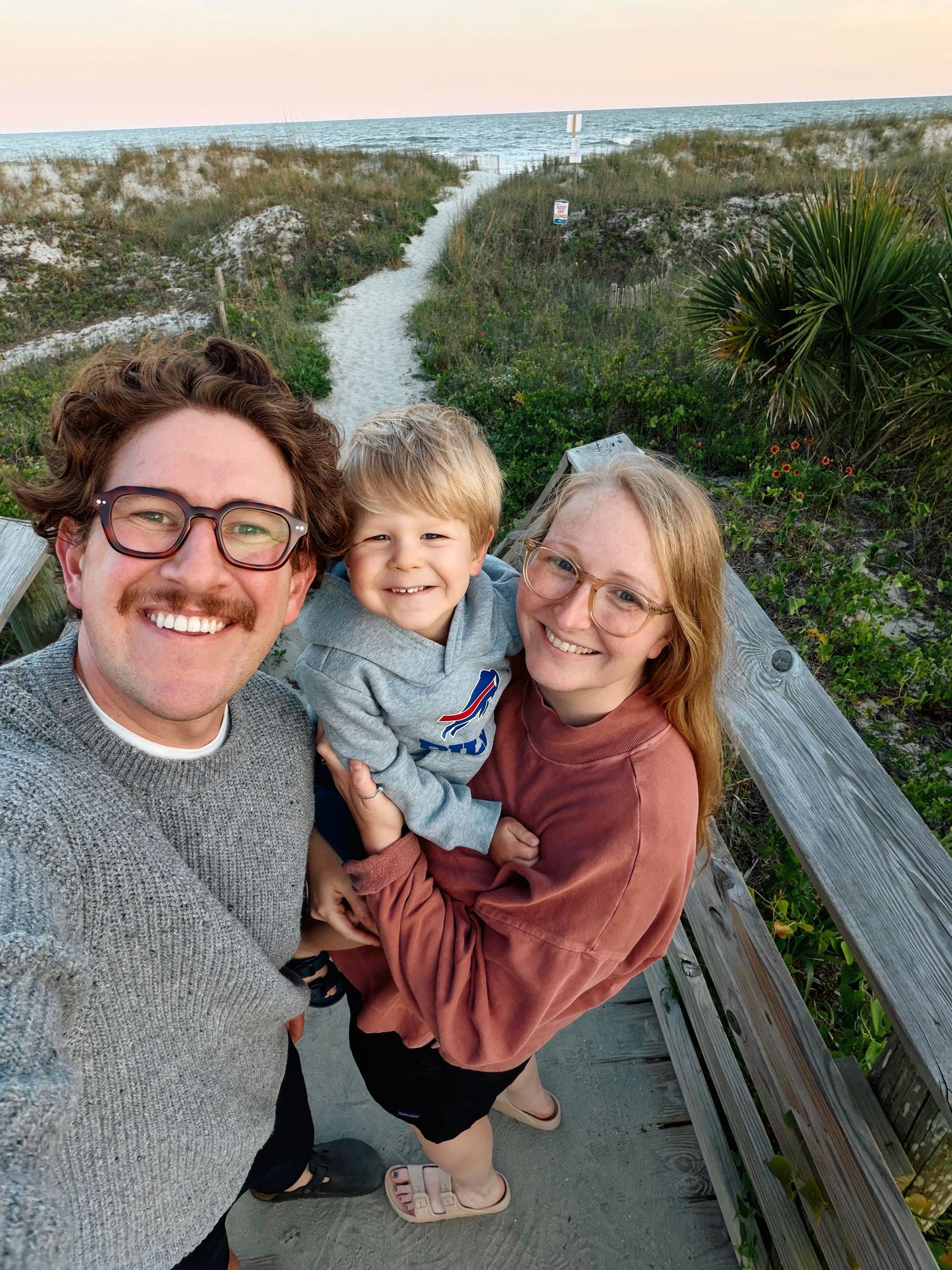 A smiling family of three poses for a selfie on a beach boardwalk at sunset, with a young child held between two adults.