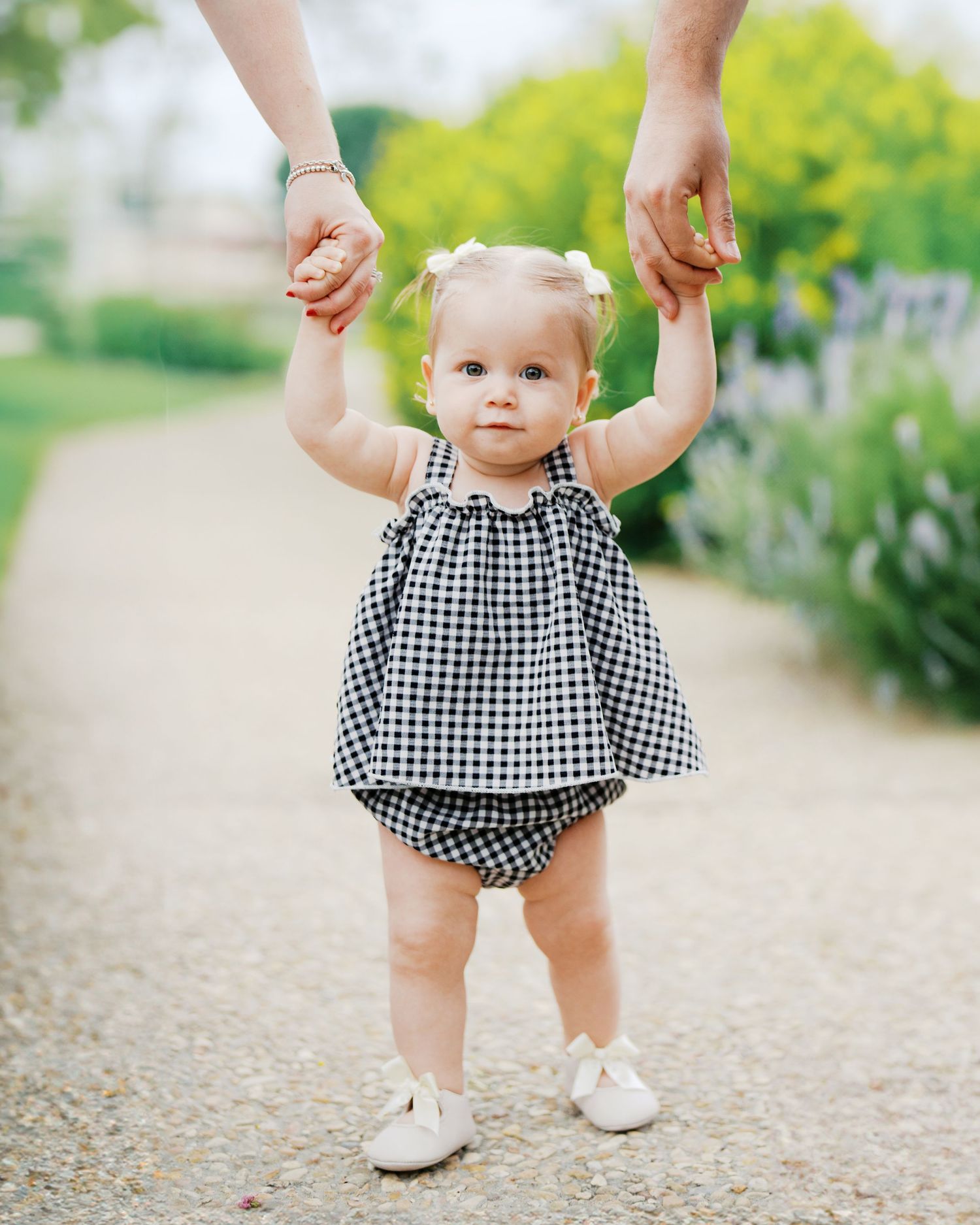 A one year old taking her first steps outdoors in San Antonio, Texas while holding her parent's hands during a milestone photo session