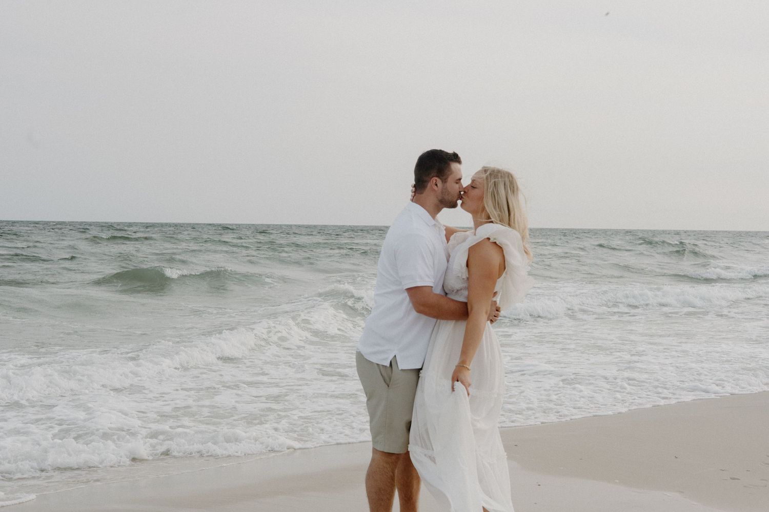 A romantic couple embracing on a beach at sunset wearing white and casual attire.
