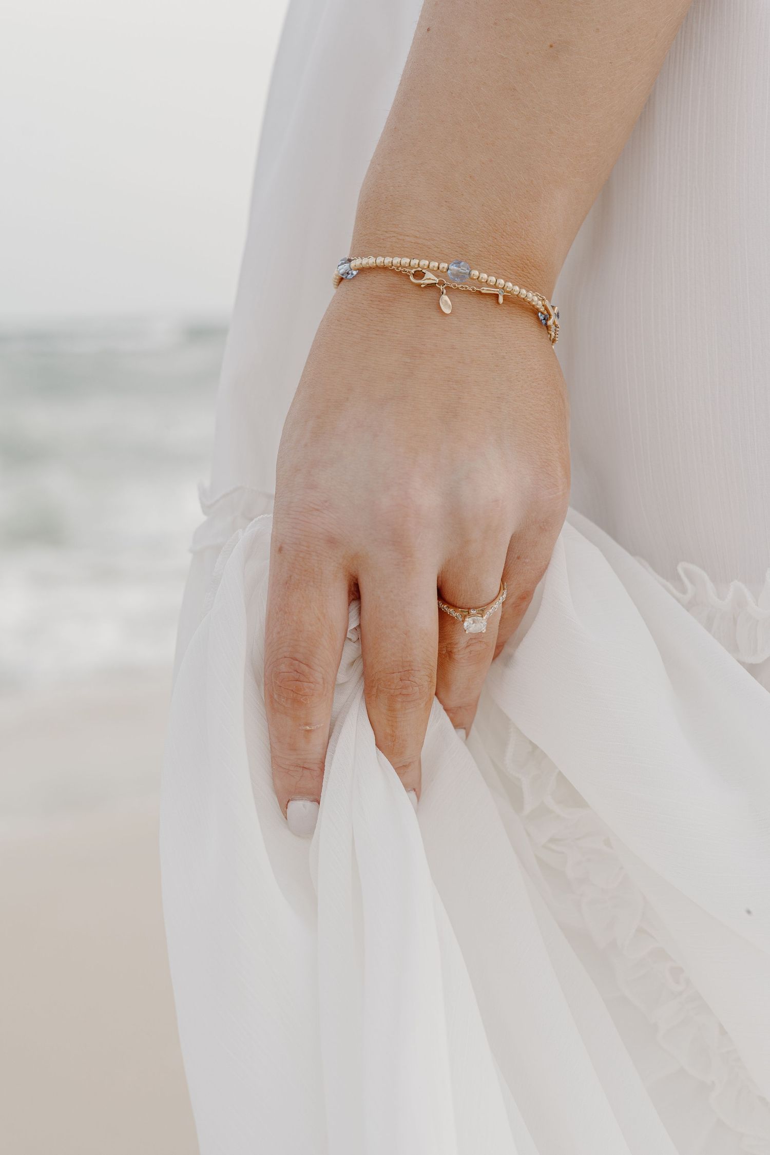 A delicate pearl bracelet and engagement ring worn against flowing white fabric at the beach.