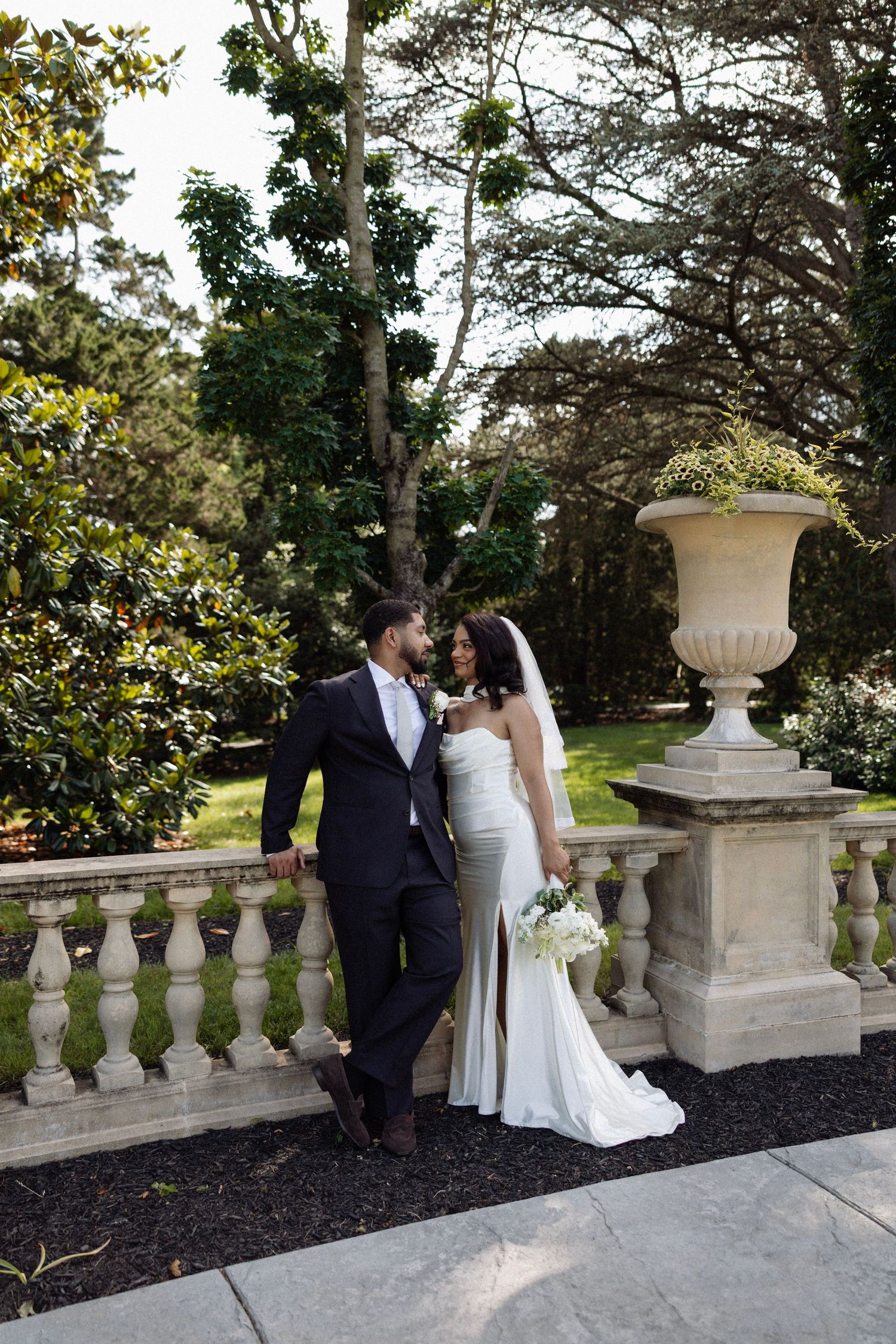 Bride and groom sharing an intimate moment on a stone balustrade in a lush garden setting.