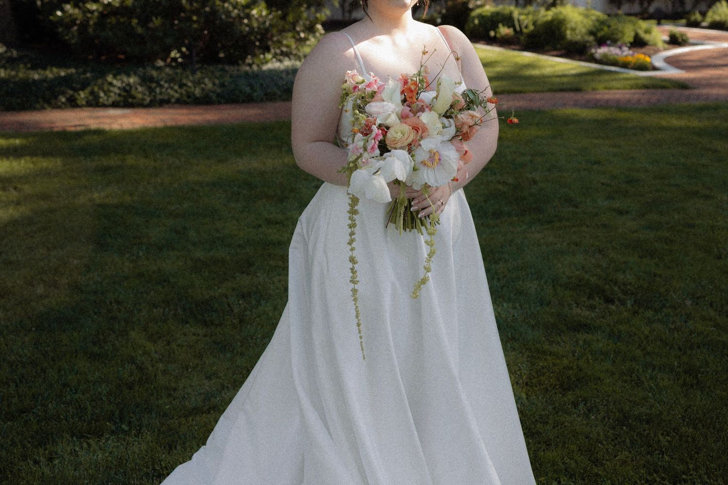 Bride in white A-line wedding gown holding peach and white floral bouquet, standing on green lawn.