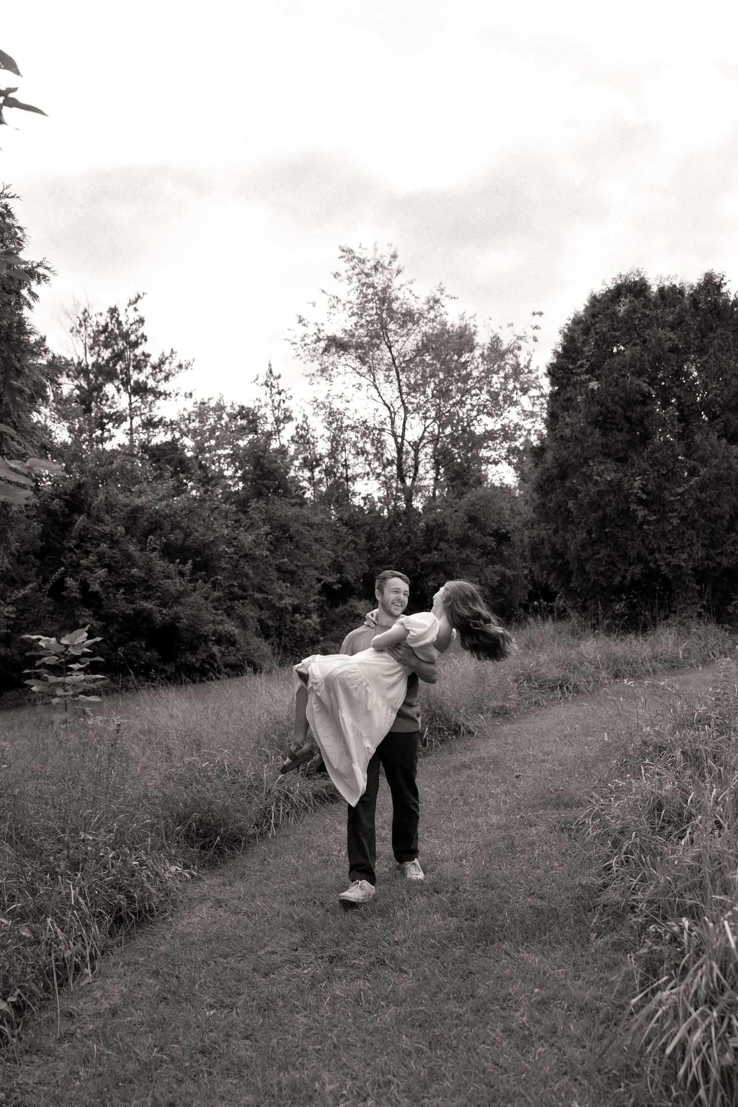 Black and white photo of a man lifting and spinning a woman on a grassy path surrounded by lush trees.