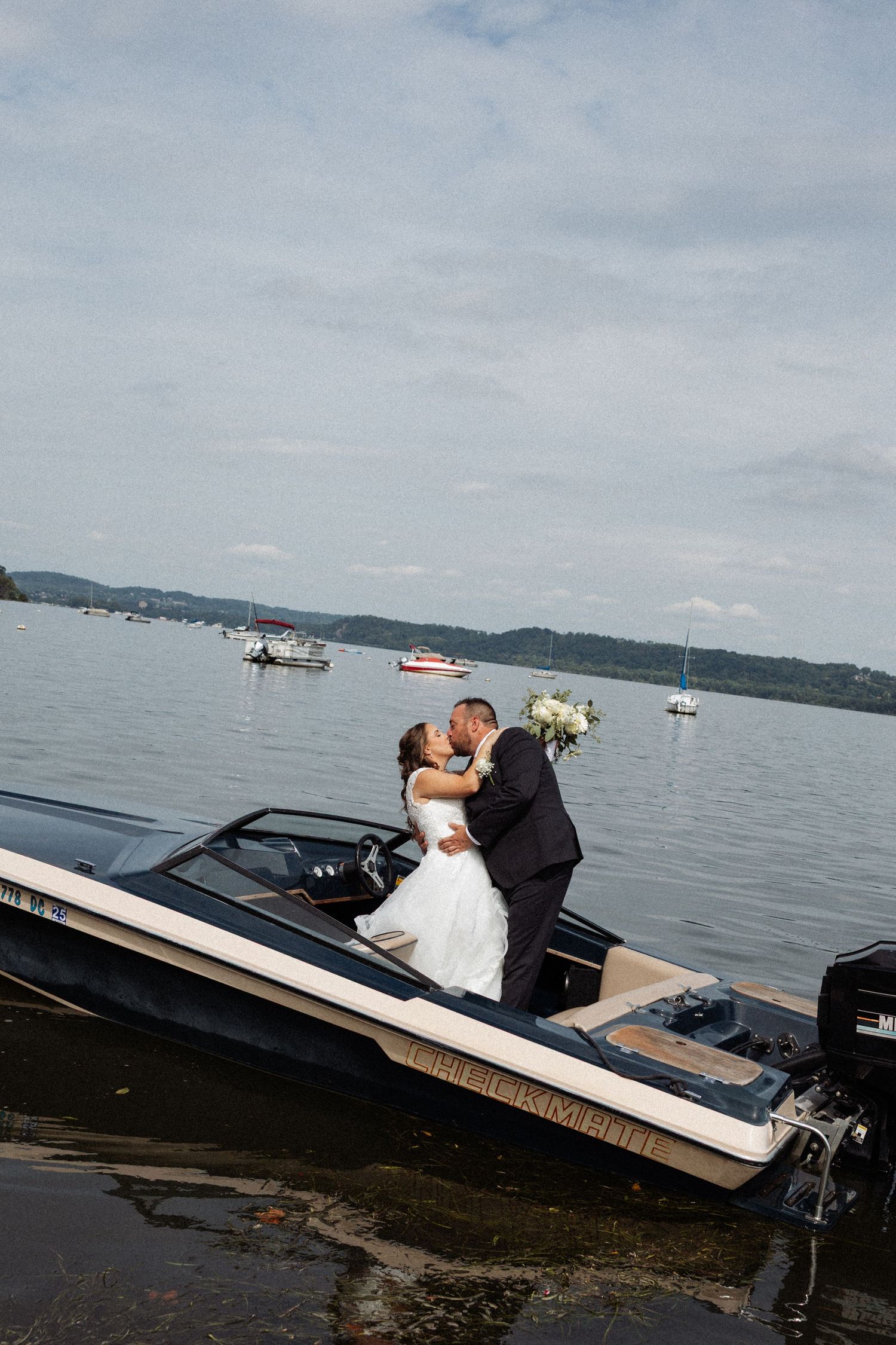 Bride and groom share a romantic kiss on a classic speedboat on a lake, with sailboats visible in the background.