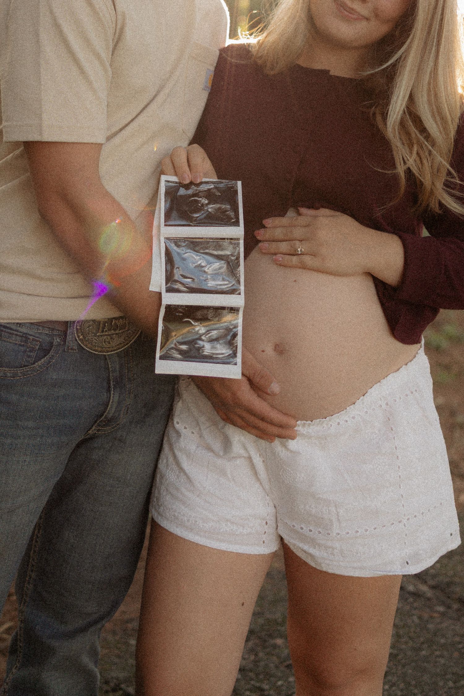 Pregnant couple holding ultrasound photos while woman cradles baby bump in white shorts outdoors.