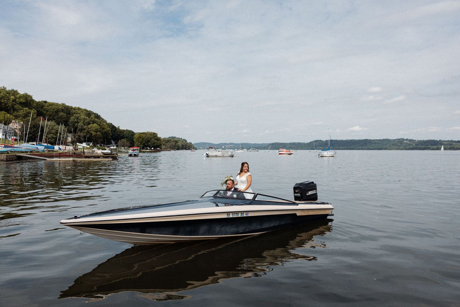 Woman driving a sleek speedboat on a calm lake with a marina and lush green hills in the background.