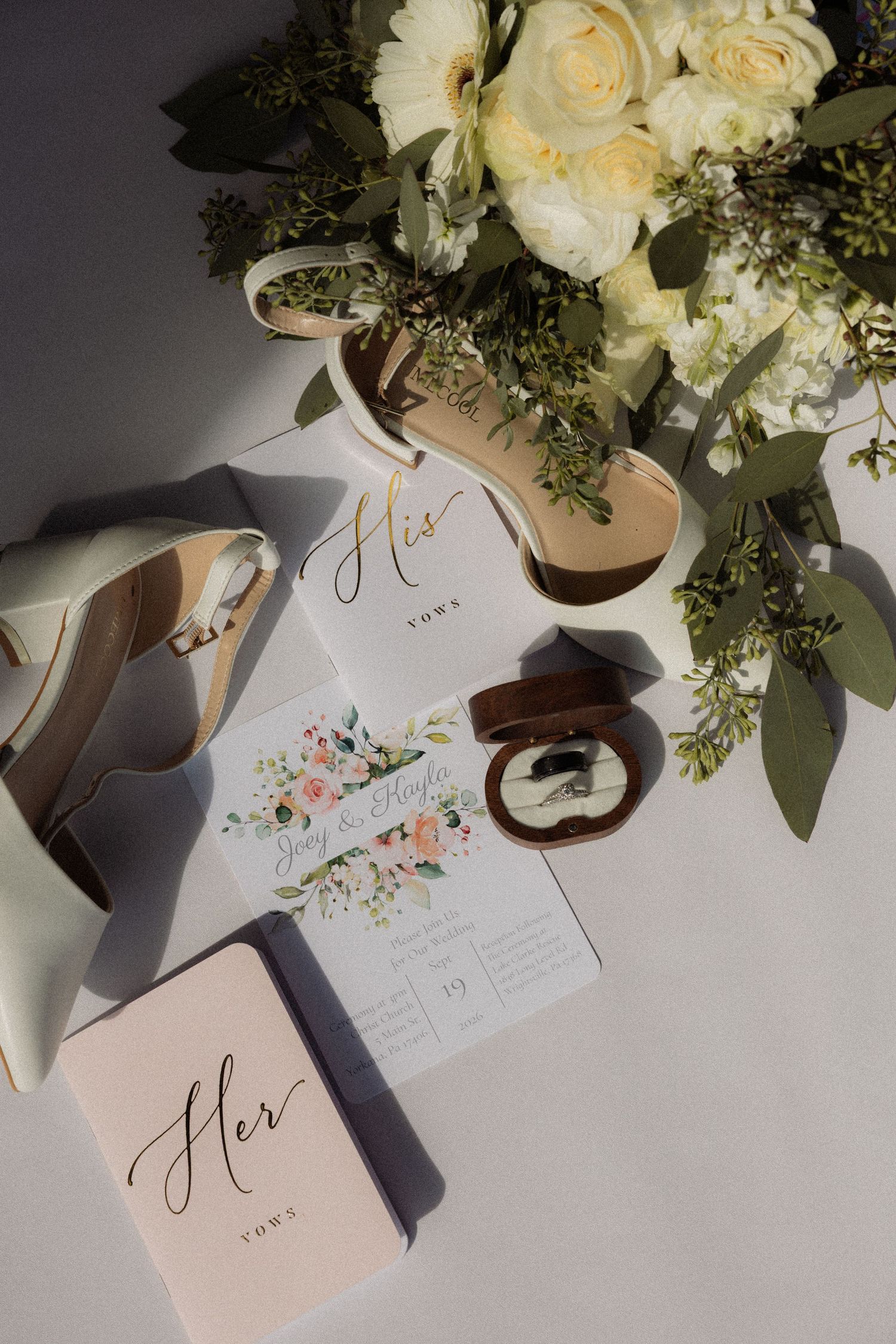 Elegant wedding flat lay with floral invitation, vow books, ring box, bridal heels, and white rose bouquet.