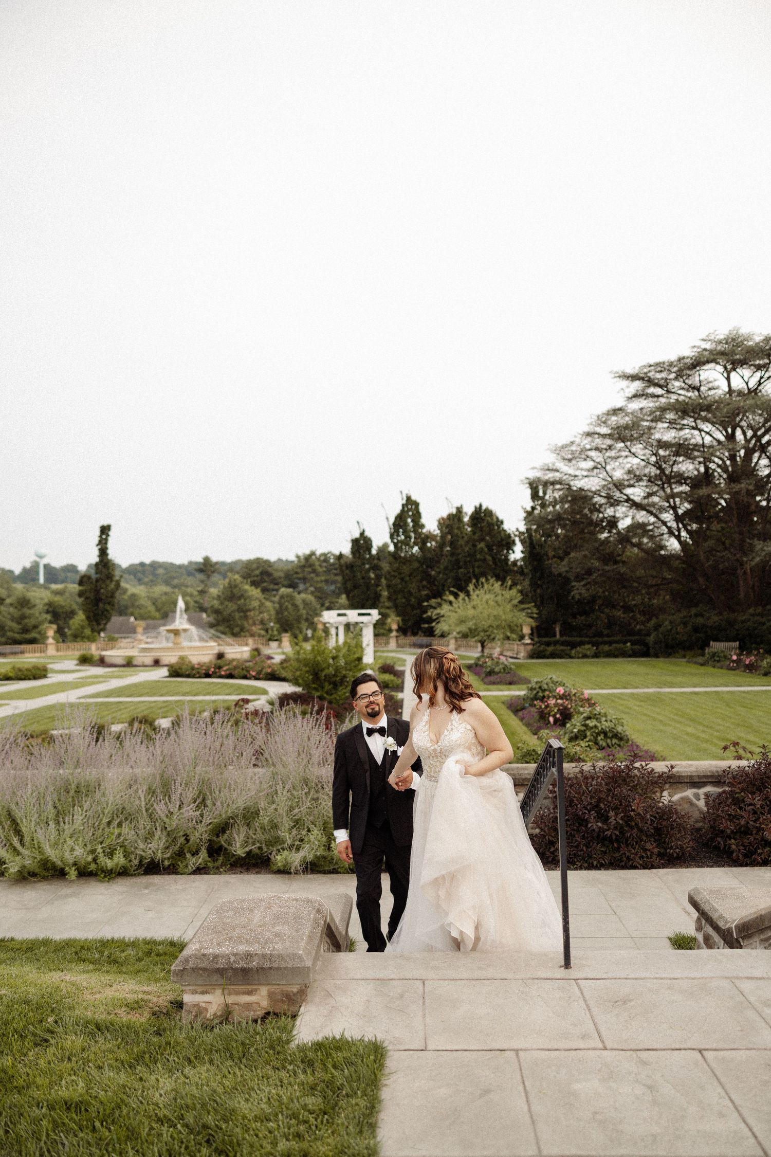 Laughing bride and groom walking up stone steps in a manicured estate garden on their wedding day.