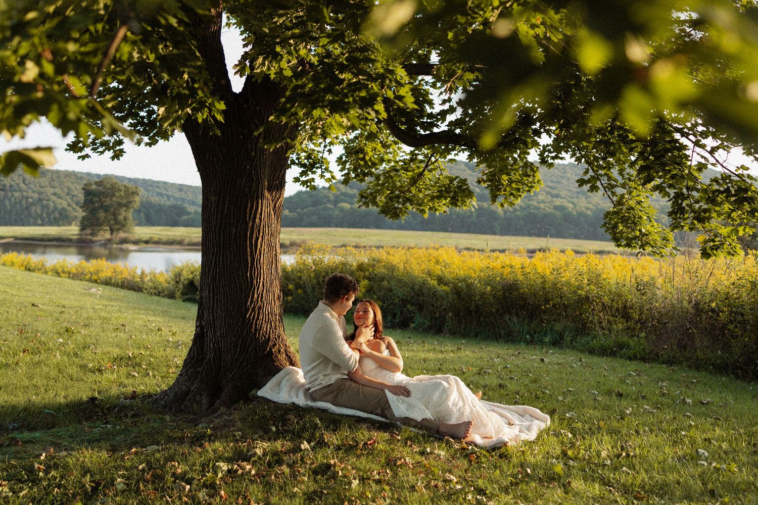 Couple sitting under a large tree on a blanket by a serene lake in golden hour sunlight.