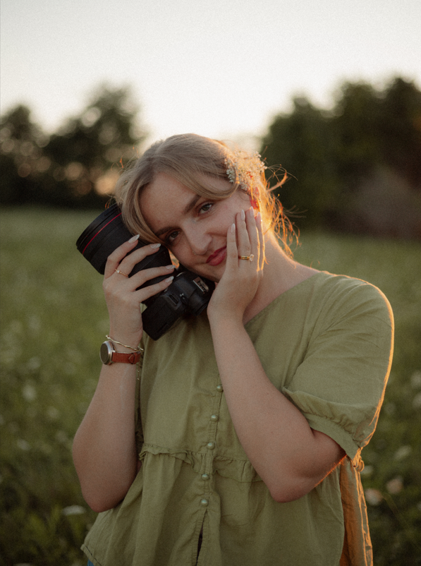 Young female photographer holding a camera against her cheek in a golden sunset field.