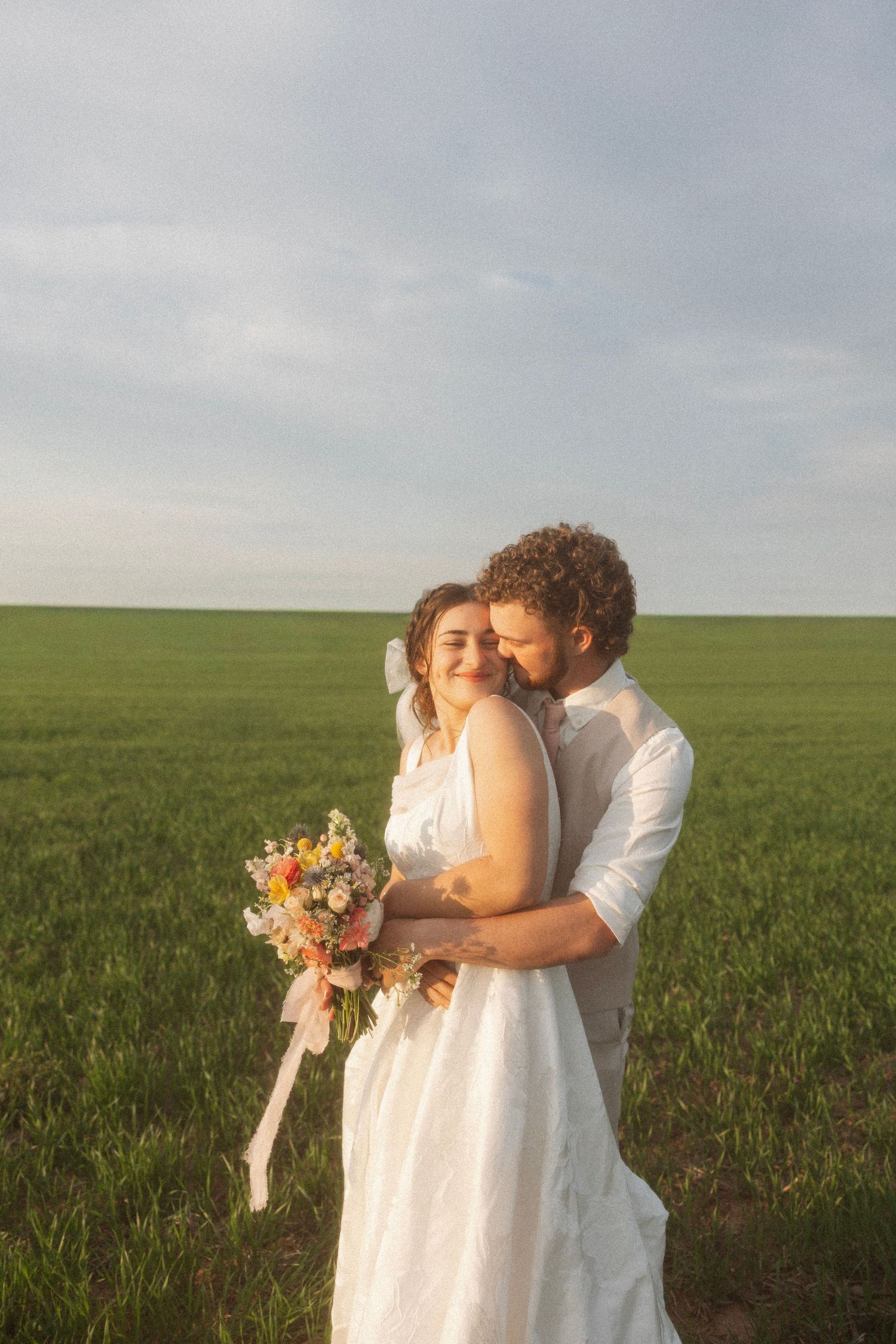 Bride and groom embracing in a green field, bride holding a colorful wildflower bouquet with pink ribbons.