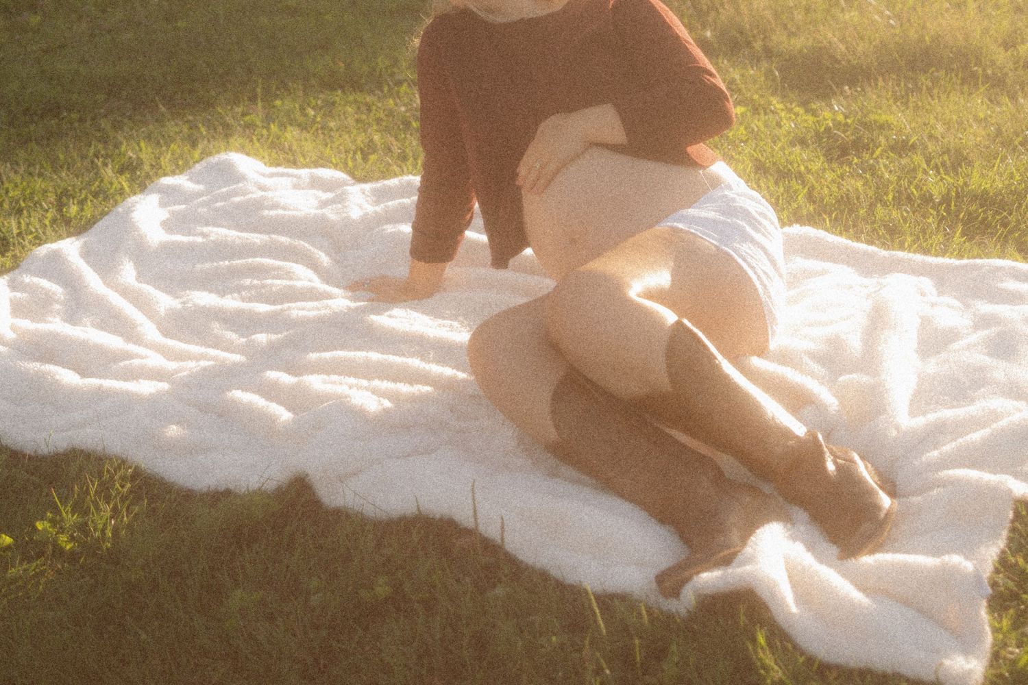 Person sitting on a fluffy white blanket in sunlit grass, wearing a rust sweater and white skirt with brown boots.