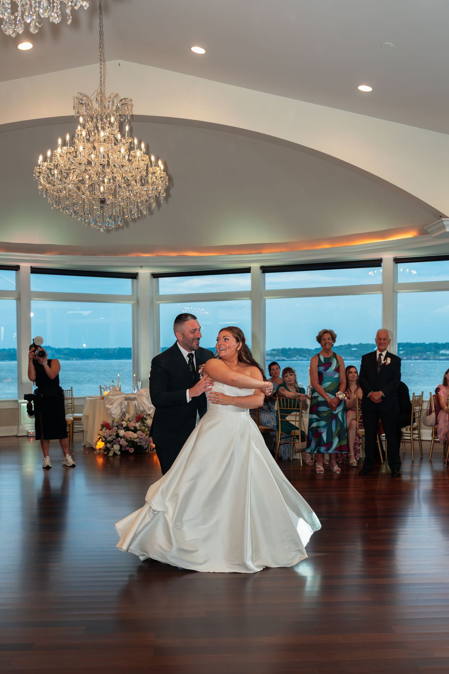 Wedding couple shares first dance under crystal chandelier in oceanfront ballroom.