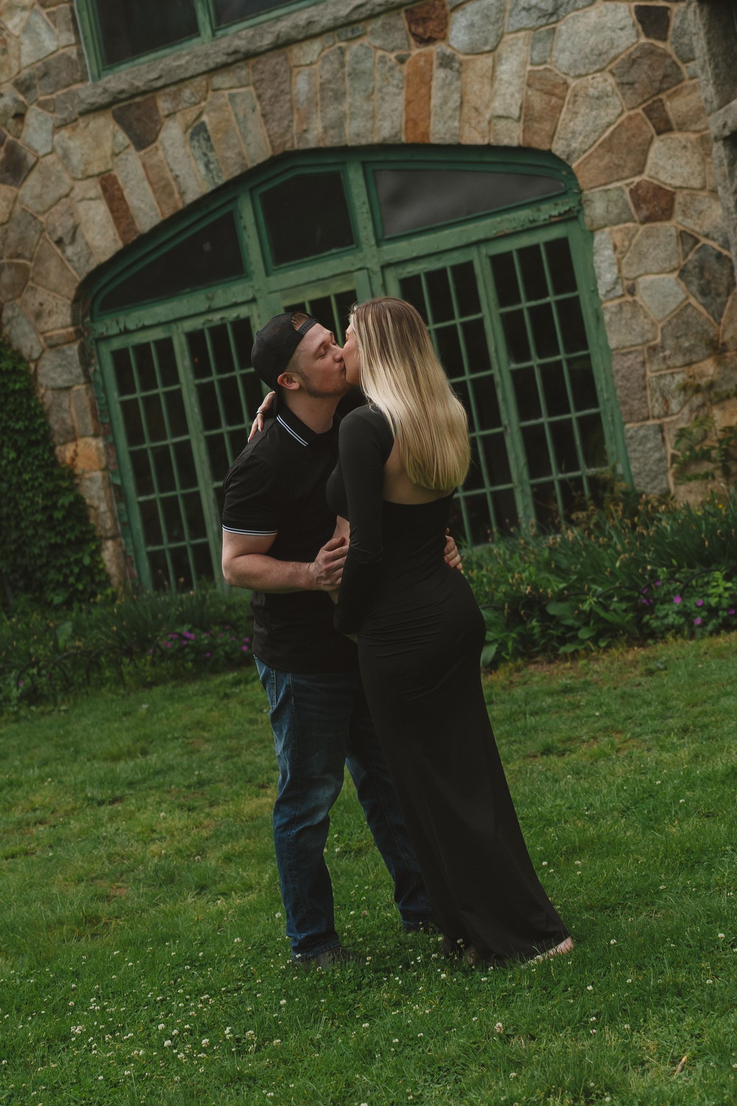 A couple shares a romantic kiss in front of a rustic stone wall with green windows and grass in the foreground.