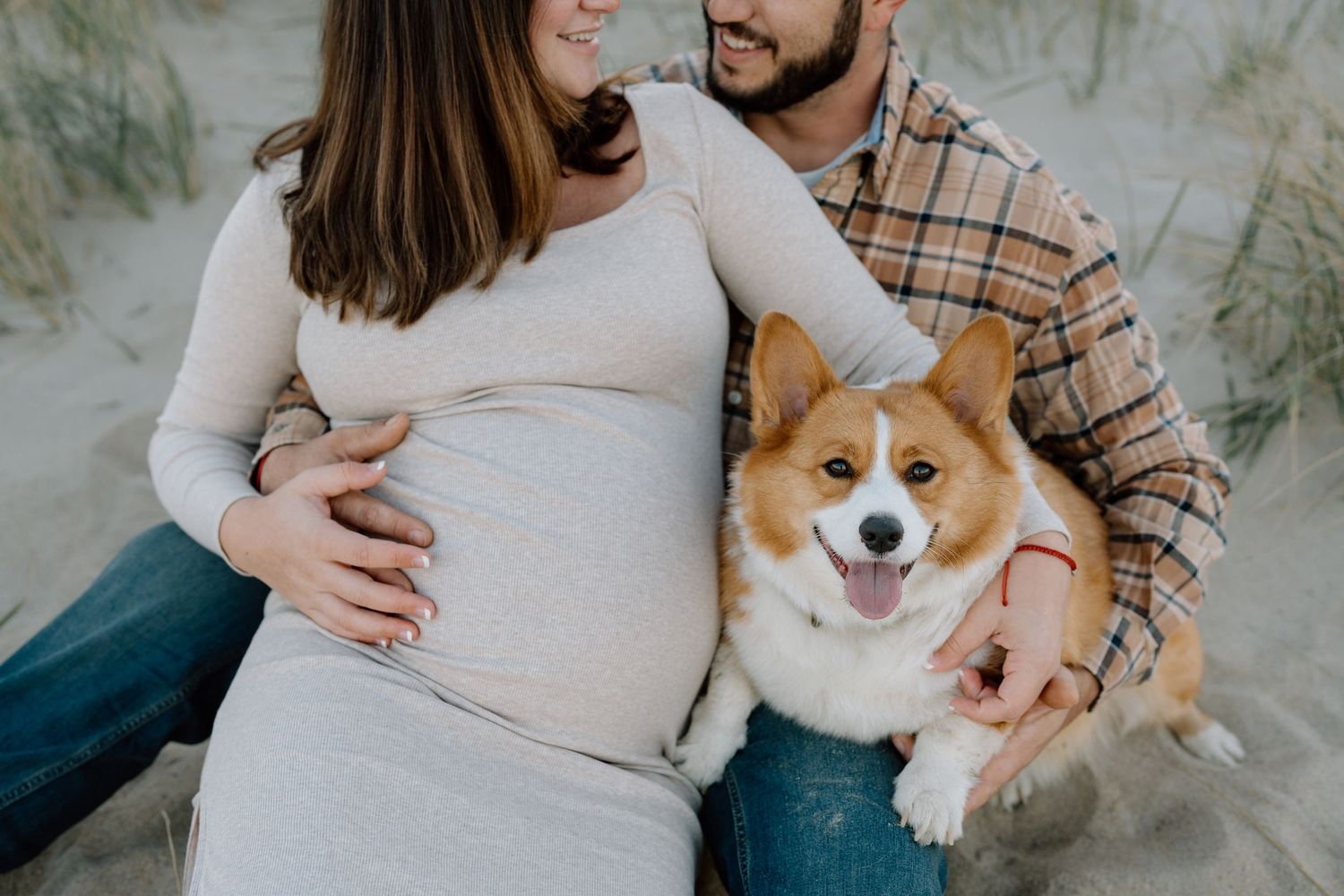 A couple poses with their smiling Corgi dog during a beach maternity photoshoot.