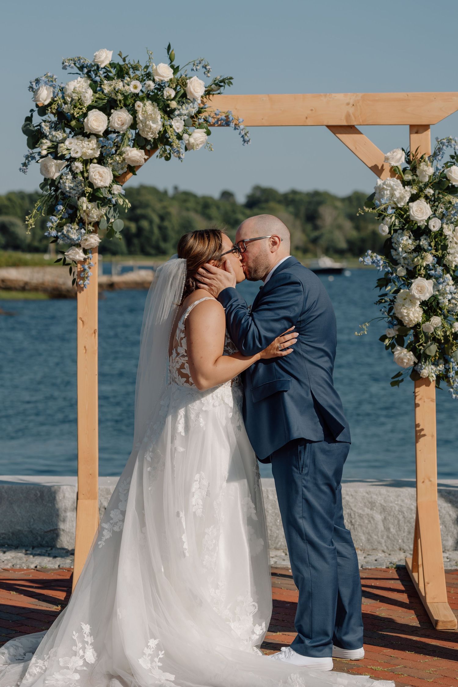Couple exchanges vows at waterfront ceremony site decorated with white floral arrangements.