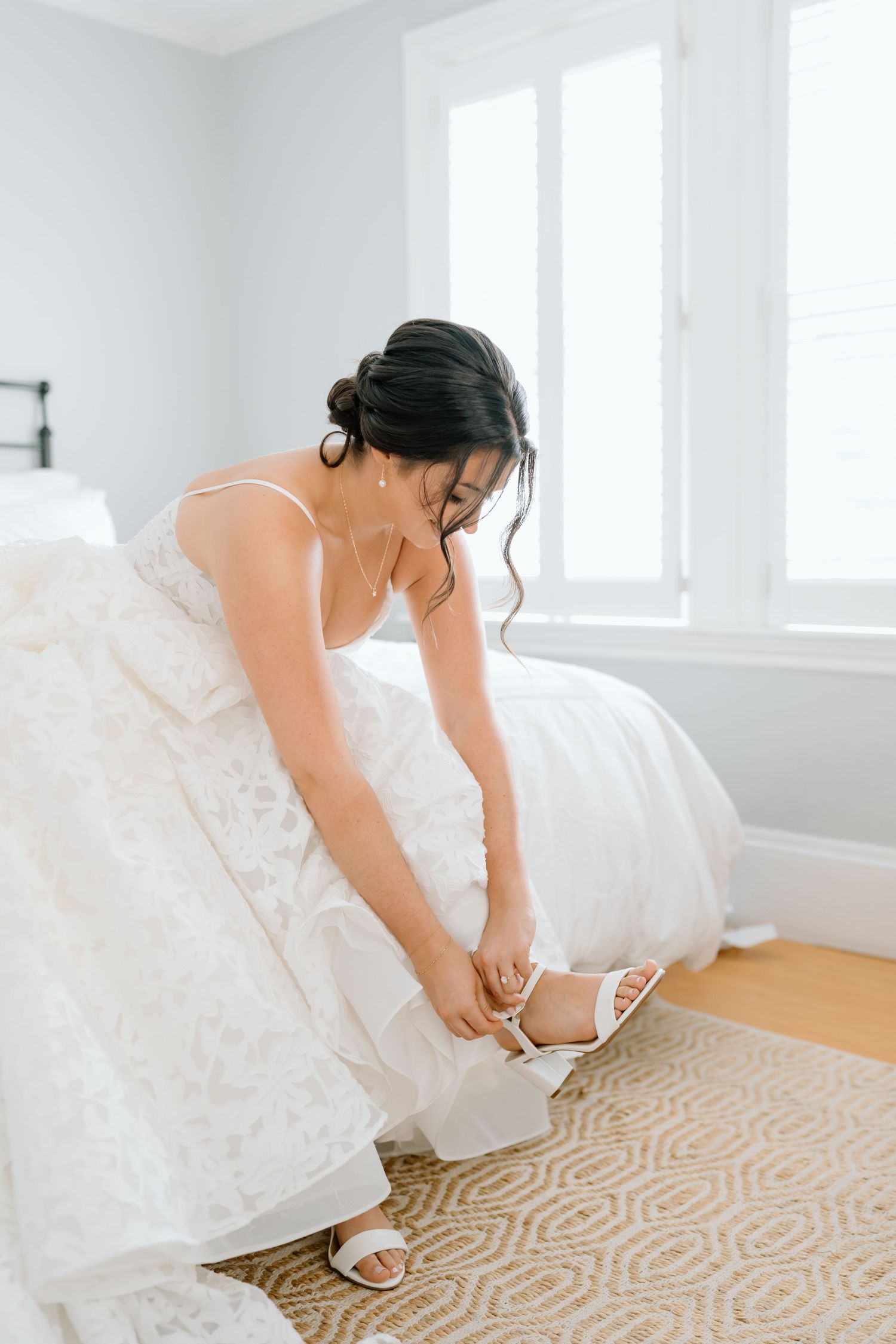 A close-up shot of a bride in a white lace dress fastening her elegant sandal on a patterned carpet.