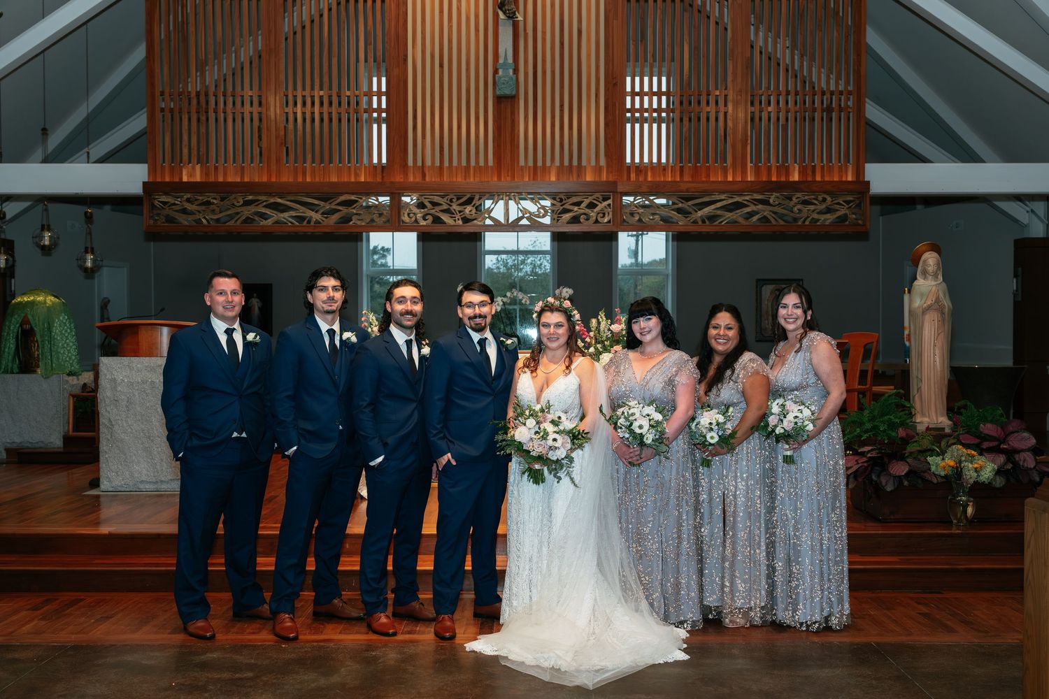 Wedding party poses in front of wooden church altar with bridesmaids in silver dresses and groomsmen in navy suits.