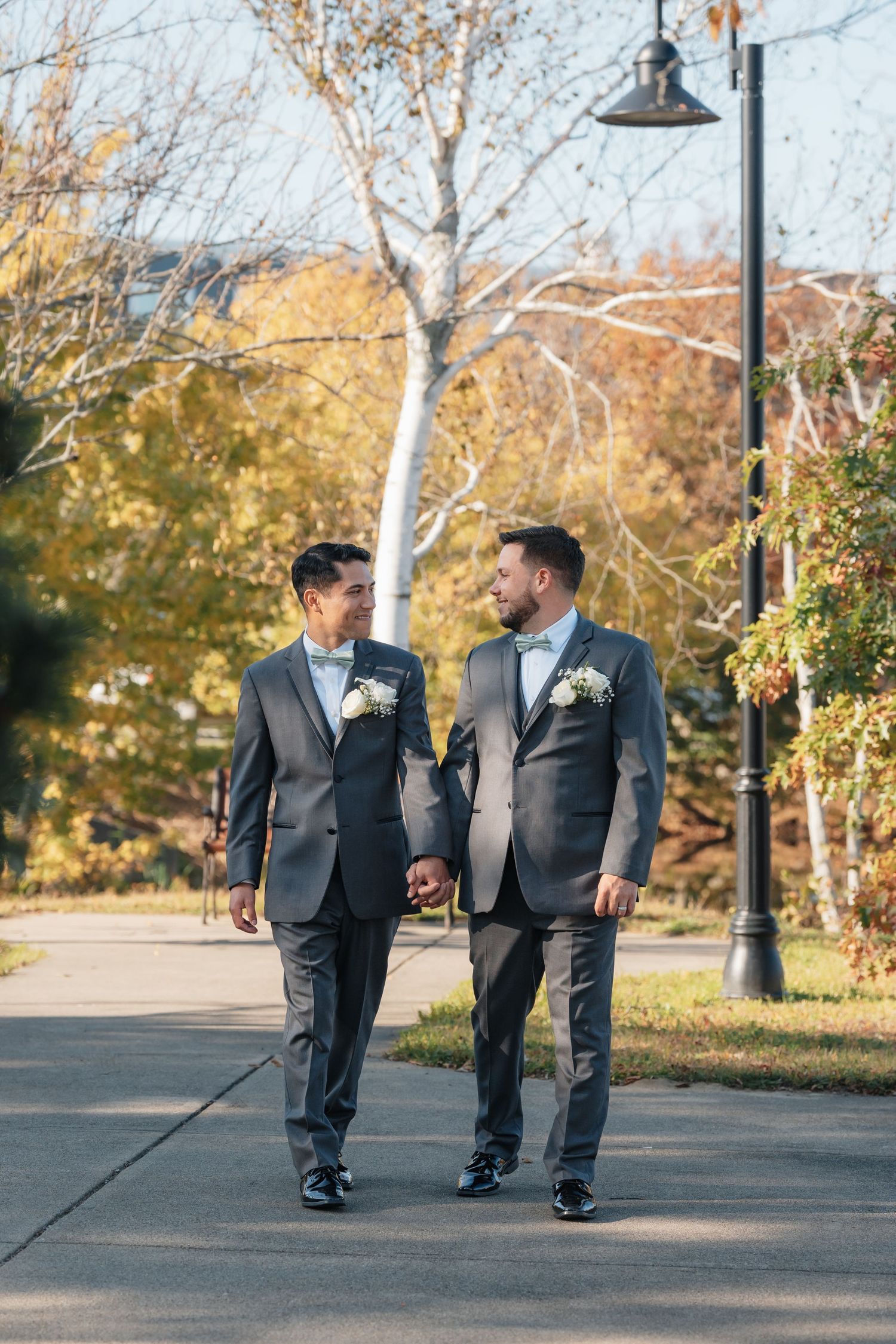 Two people in dark suits walk together on a path with fall trees and lampposts.