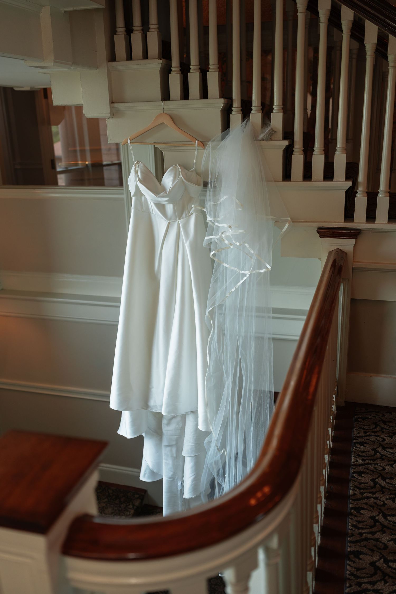 White wedding dress and veil hanging on wooden staircase with classic banister.