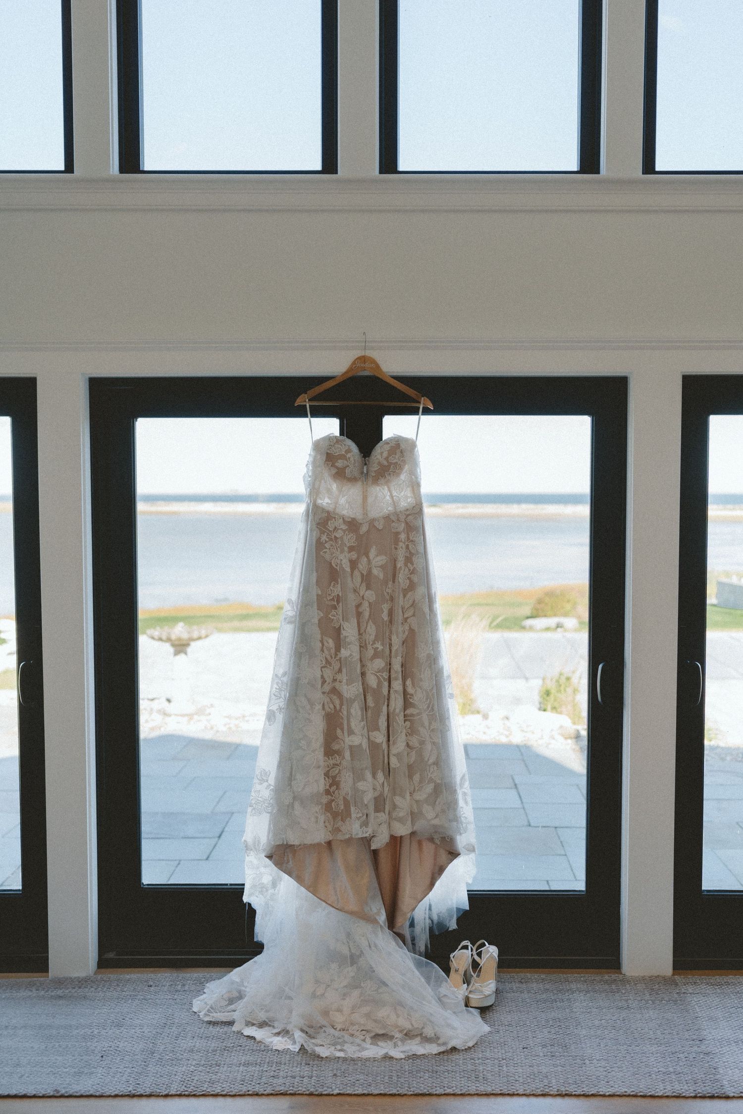 Lace wedding dress hanging in front of large windows overlooking a coastal view.