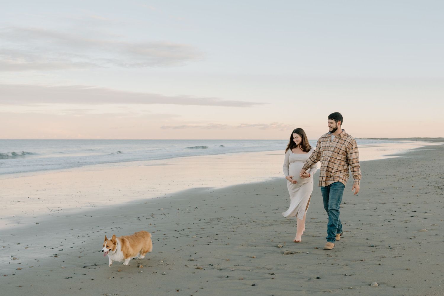 A couple walks along the beach with their Corgi dog during sunset.