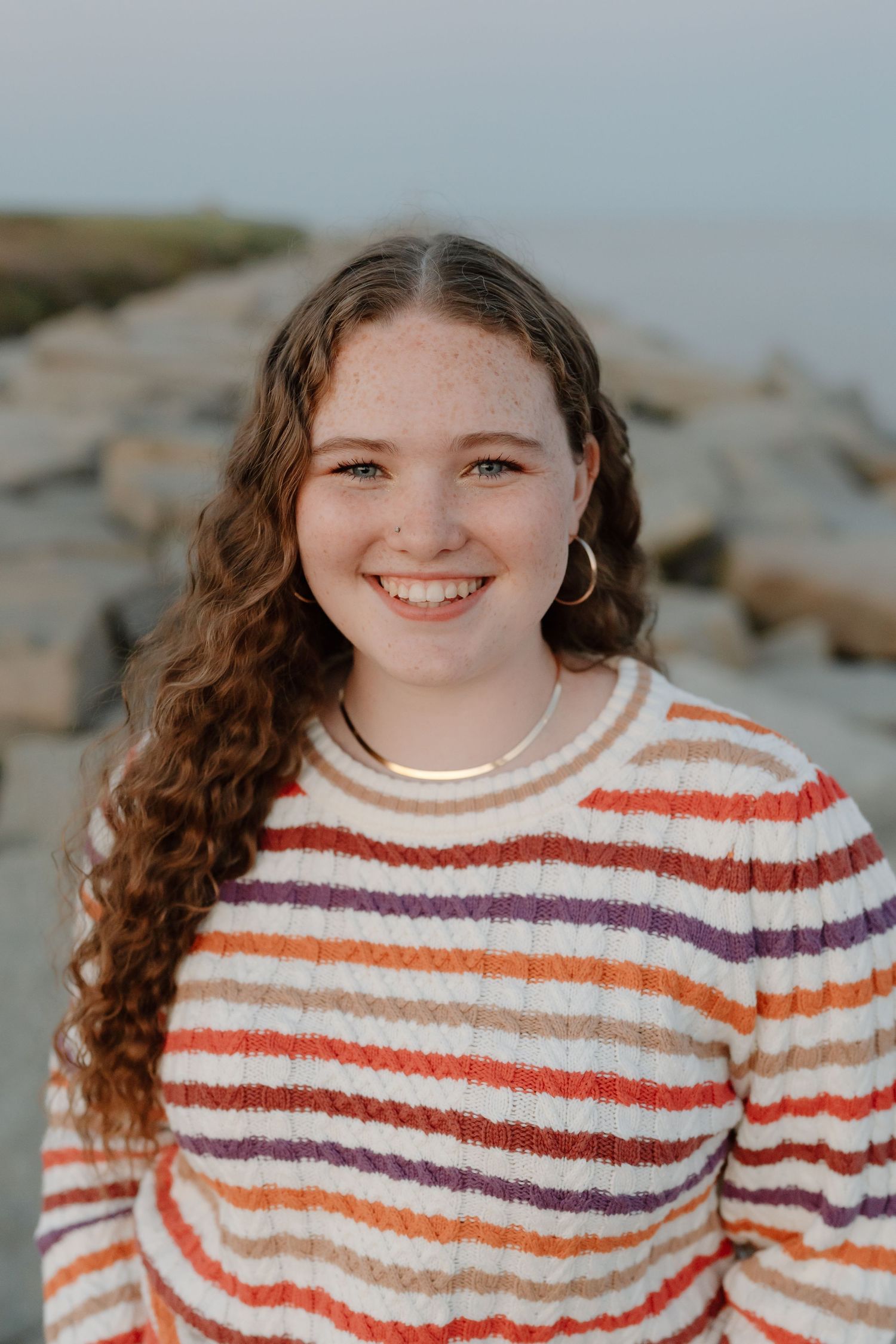Coastal portrait featuring someone in multicolored striped sweater against rocky beach background.