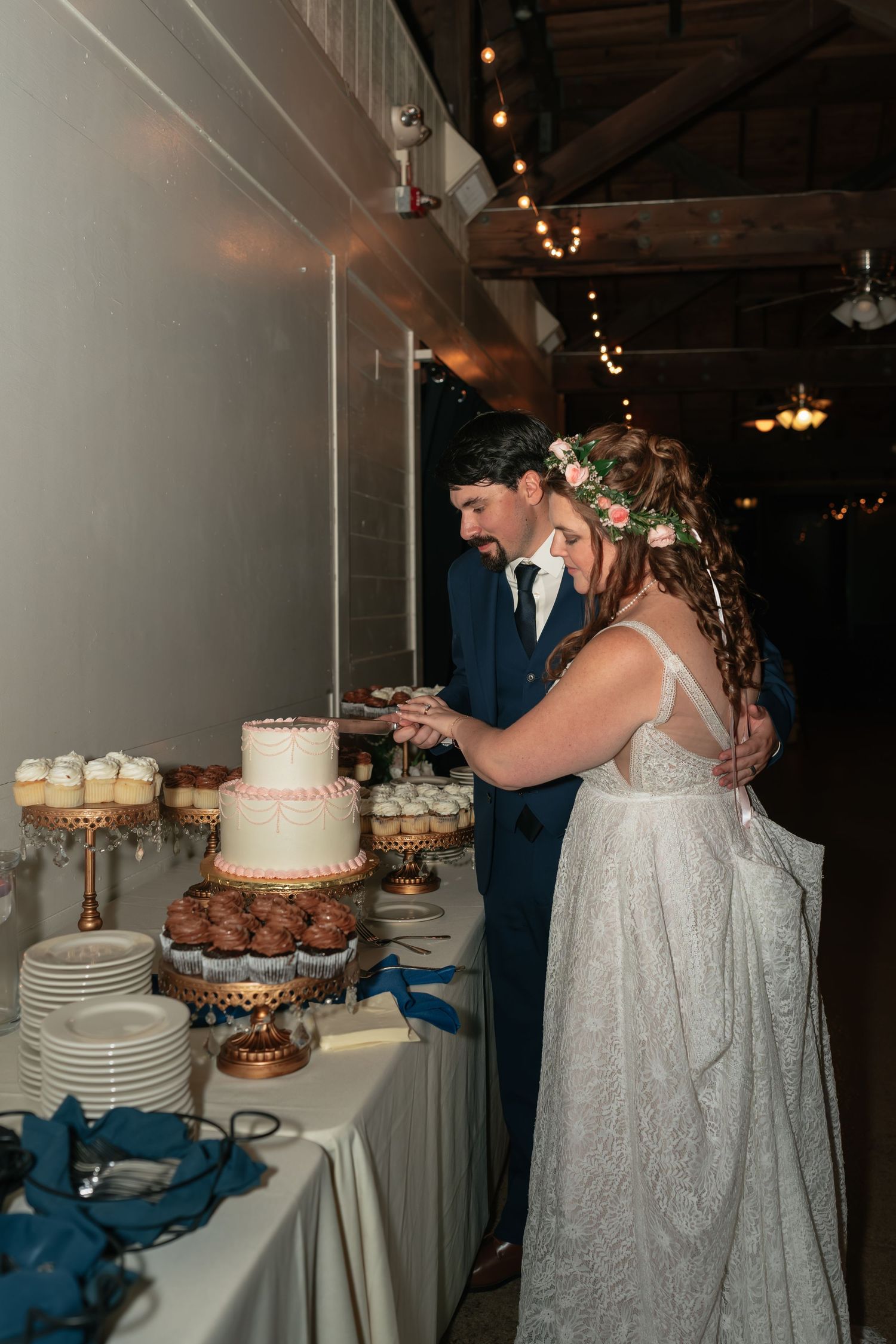 Couple serving wedding cake at dessert table decorated with cupcakes and string lights.