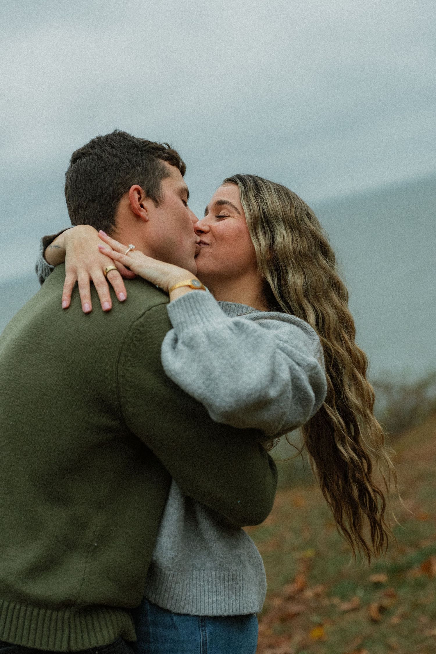 A couple in casual sweaters shares a passionate kiss against a cloudy coastal backdrop.